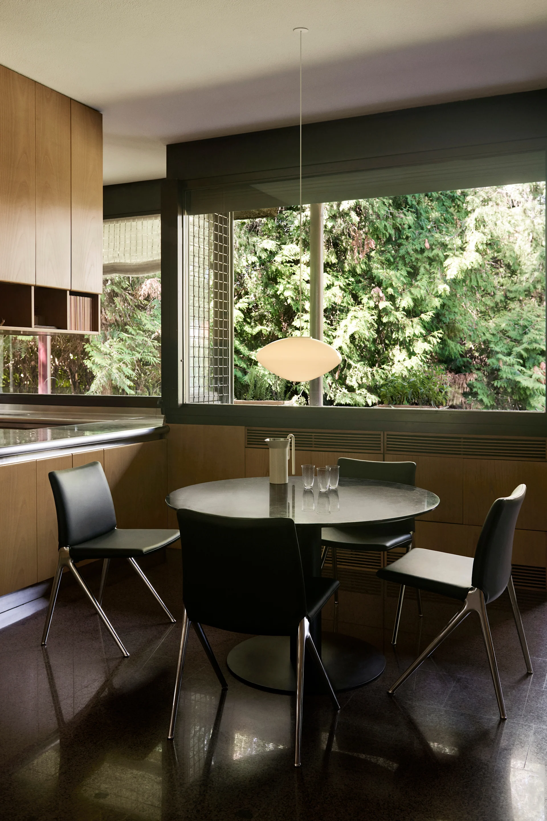 A modern dining area featuring a round black table, four black chairs, and a white oval pendant light. Light wood cabinets line one wall, with large windows overlooking green trees.