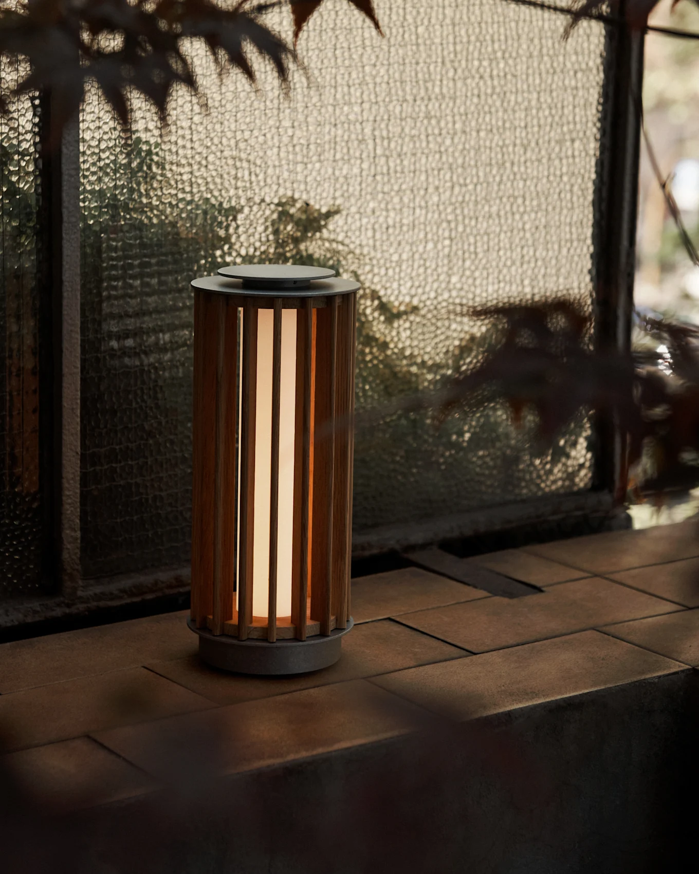 A cylindrical portable lamp with a slatted wooden body and warm glow, on a tiled surface by a textured glass window.