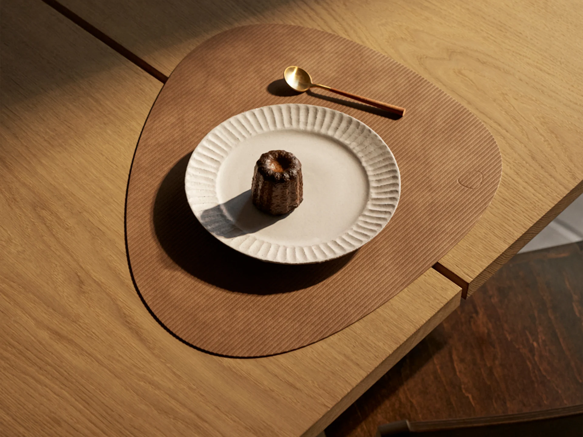 A canelé pastry on a ribbed plate, a gold spoon, and a triangular placemat on a wooden table, lit by sunlight.