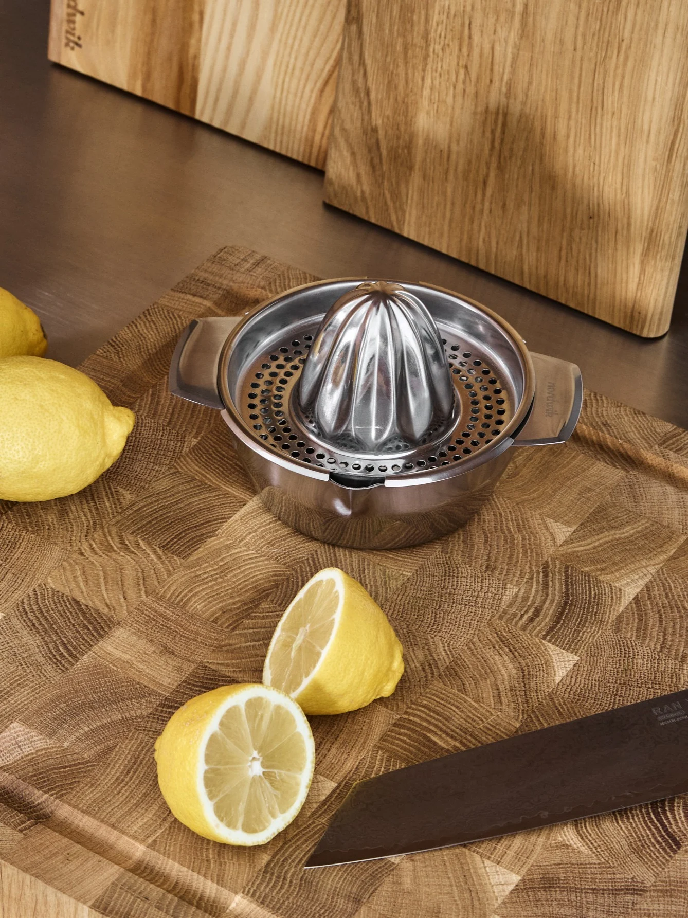Stainless steel manual juicer, whole and halved lemons, and a knife on a wooden cutting board.