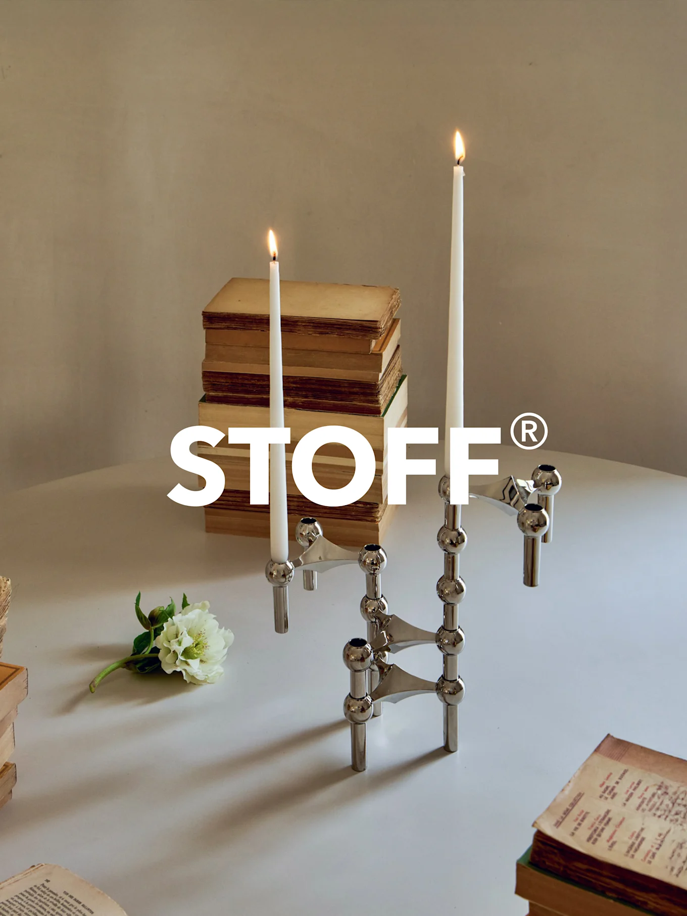 Polished chrome modular candelabra with two lit candles, surrounded by old books and a white flower on a table. STOFF® logo.