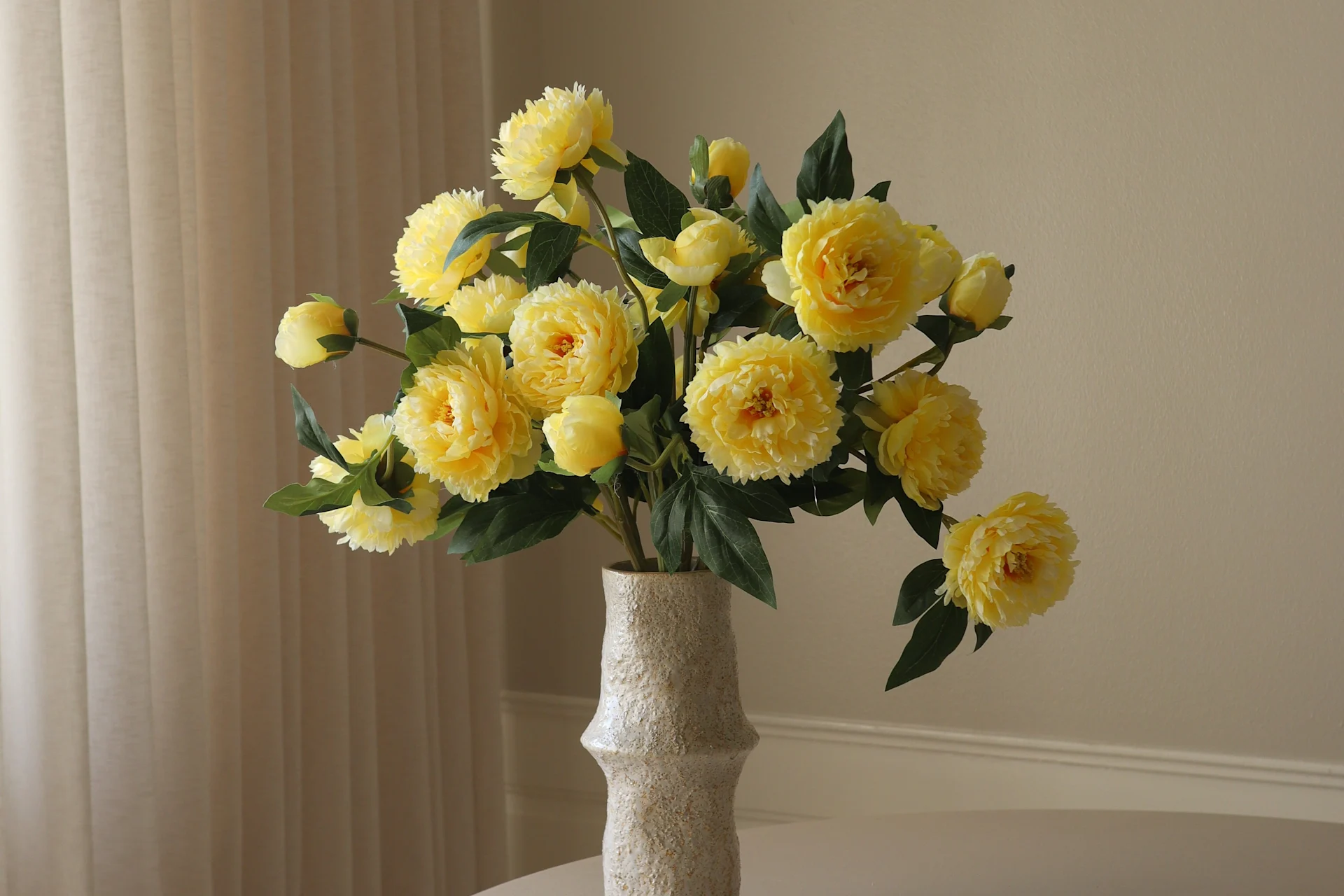 A light-colored, textured vase holds a vibrant bouquet of yellow peony-like flowers and green leaves against a light background.