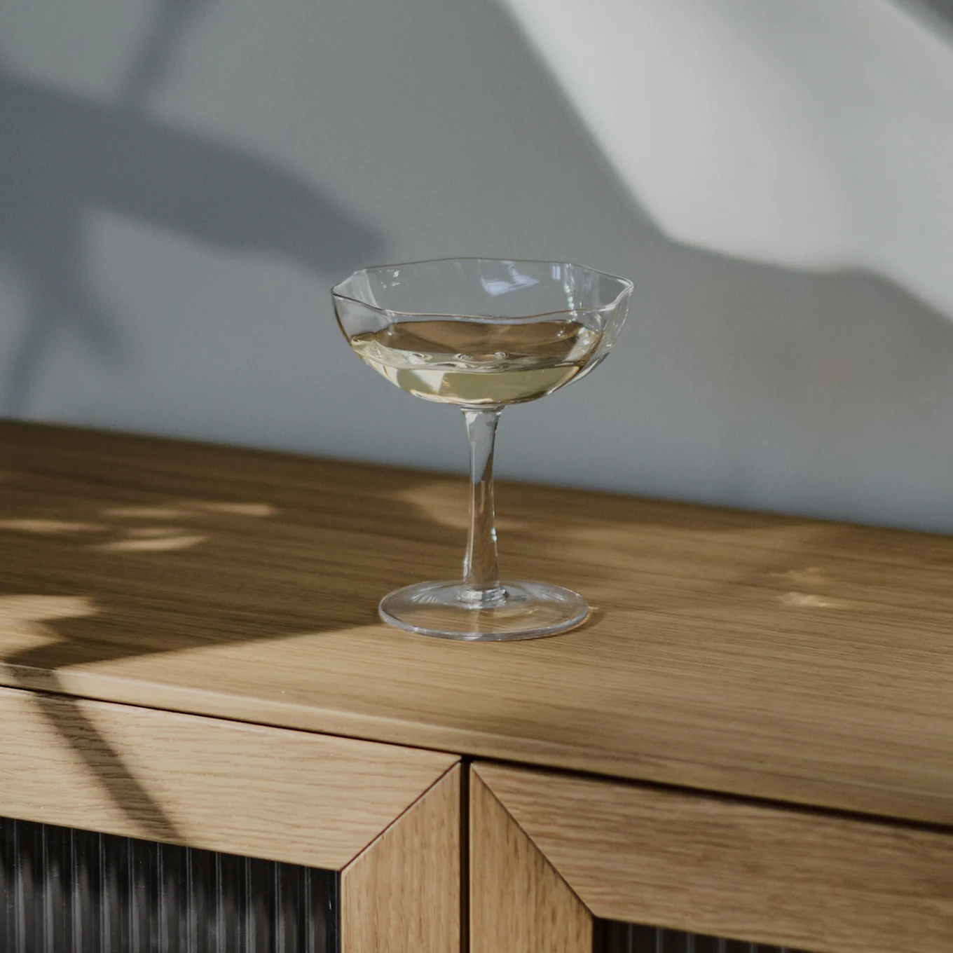 A clear coupe glass filled with a light-colored drink sits on a light brown wooden cabinet, with shadows cast on the wall behind.