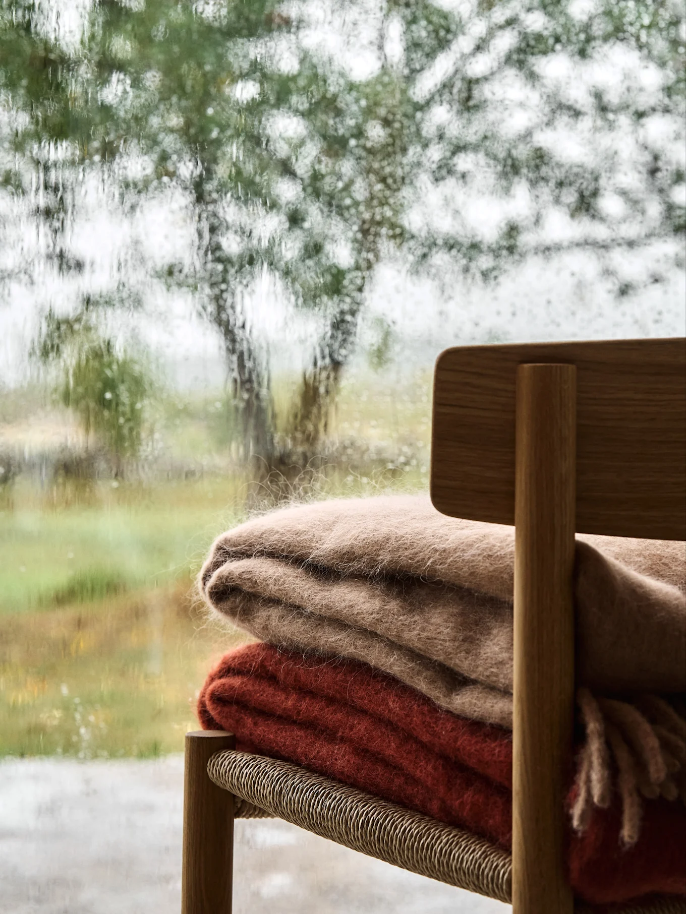 Cozy scene with two fluffy blankets on a wooden chair, seen through a rain-streaked window overlooking a blurry green landscape.