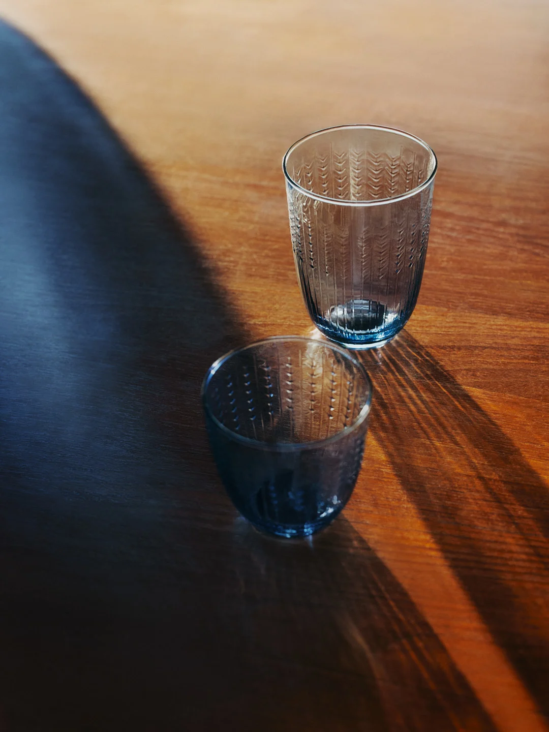 Two blue-tinted, textured glass tumblers on a wooden table, with long shadows cast across the surface.