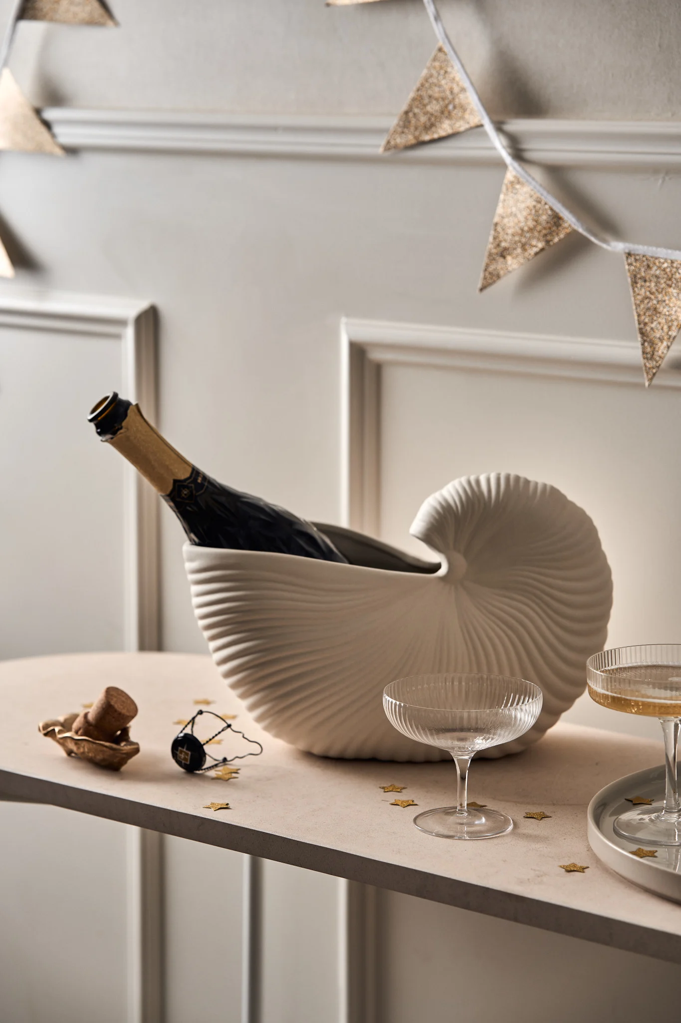 Discover how to throw a party with a drinks station, here you see the a bottle of champagne chilling in the Ferm Living shell vase with some champagne coupes arranged on a table as a self service drink station. 