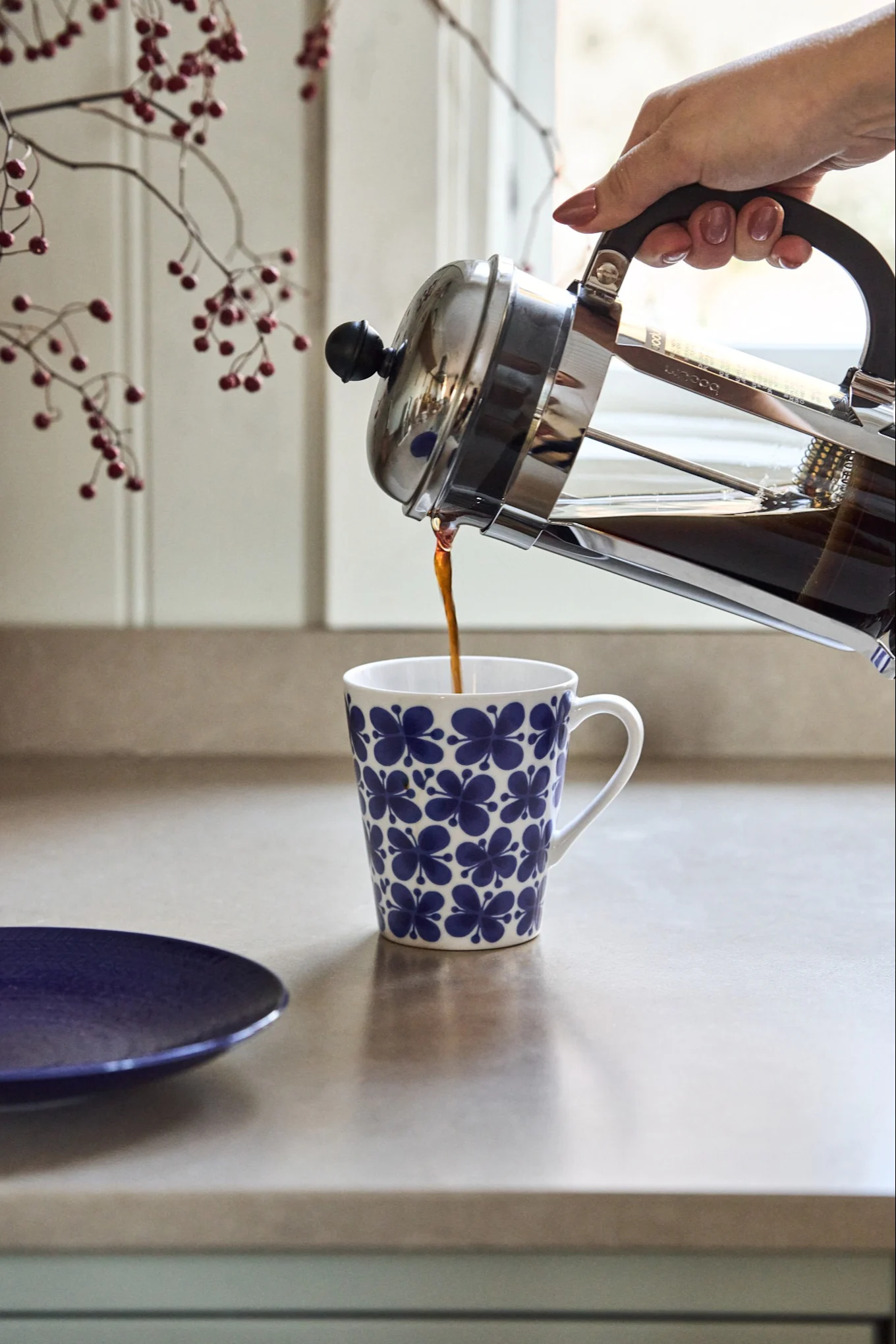A hand pours coffee from a French press into a white mug with a blue floral pattern on a kitchen counter.