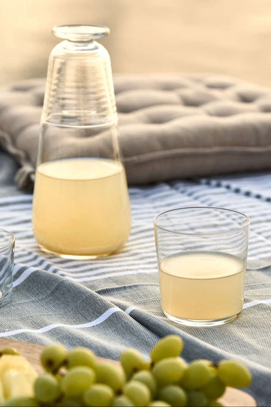 Discover simple summer recipes: here you see a bottle of lemon juice with a glass and a bunch of grapes laid out on a picnic table. 