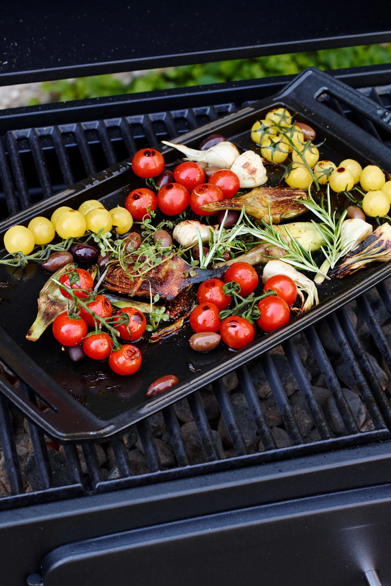 Grilled vegetables and herbs on a black tray: red and yellow cherry tomatoes, artichokes, garlic, olives, rosemary, and thyme.