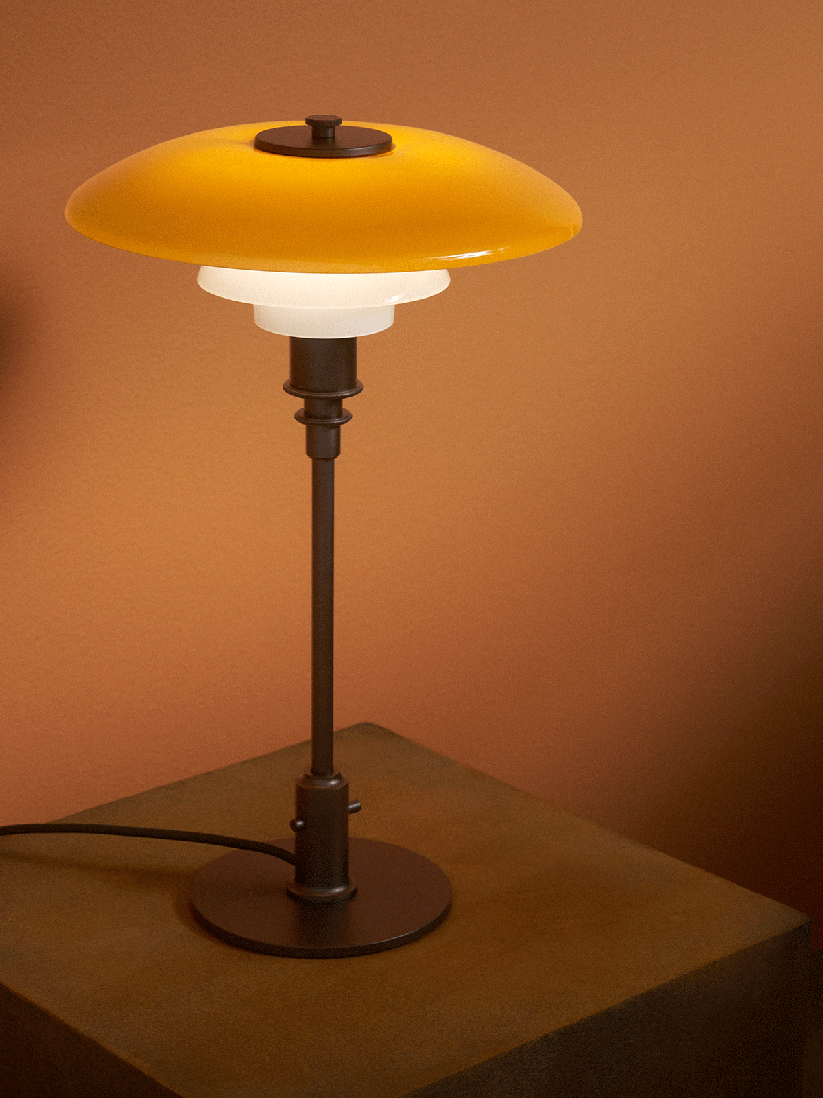 A modern table lamp with a yellow top shade and two white shades sits on a richly textured wooden pedestal against a brown wall.