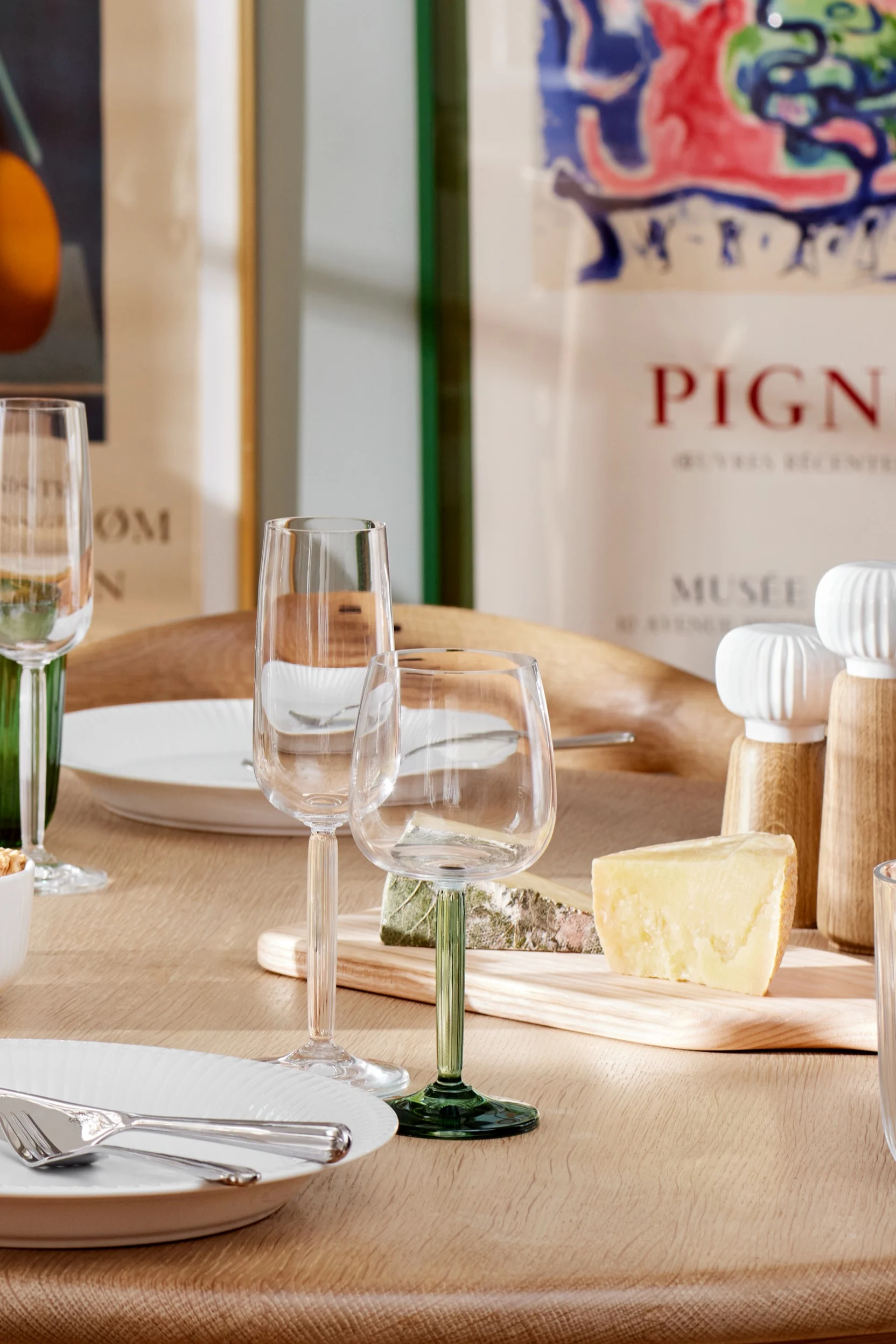 Hammshoi wine glasses with clear and green stems stand on a dining table in the sun.