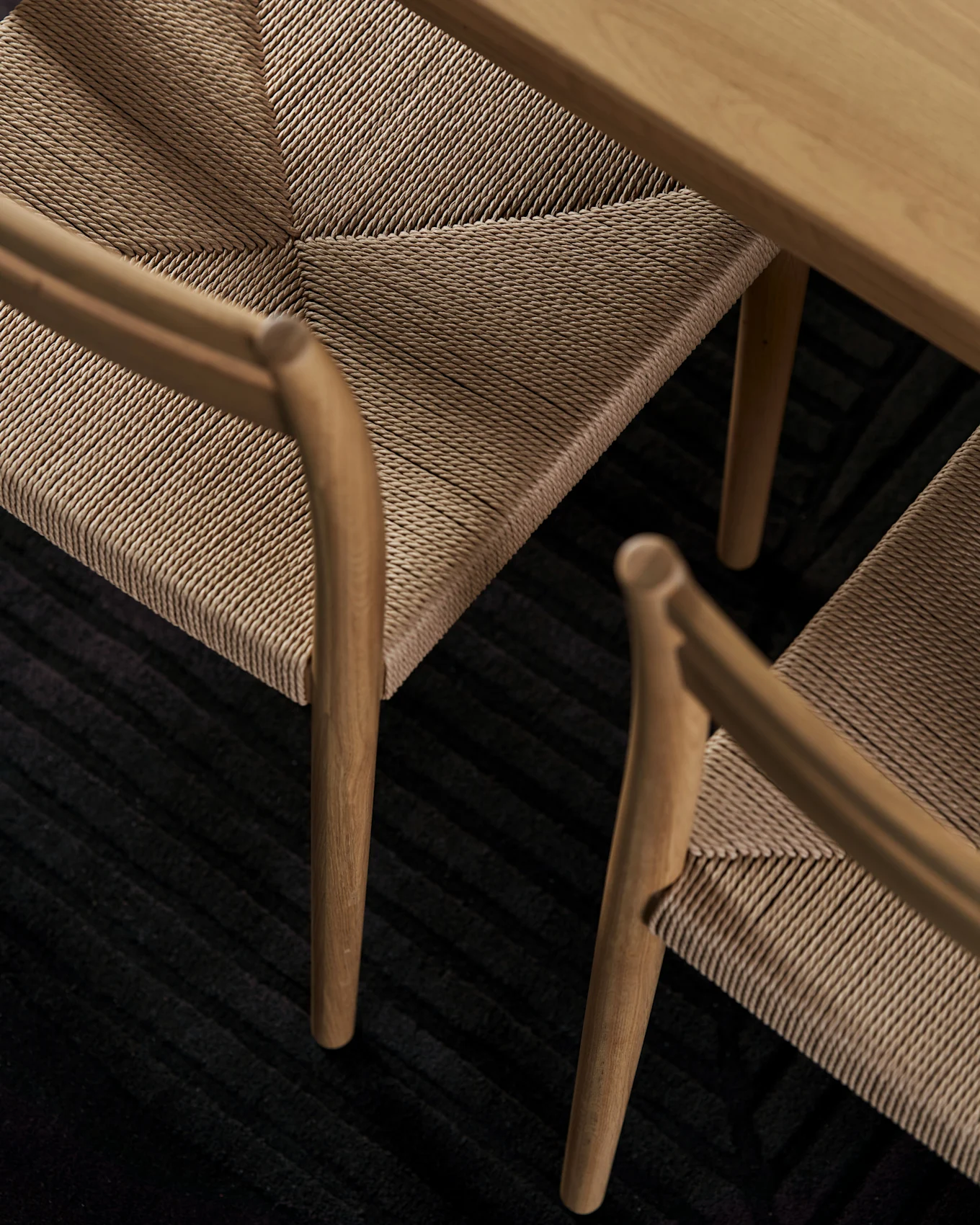 Close-up of wooden chairs with woven rope seats under a wooden table on a dark ribbed rug.