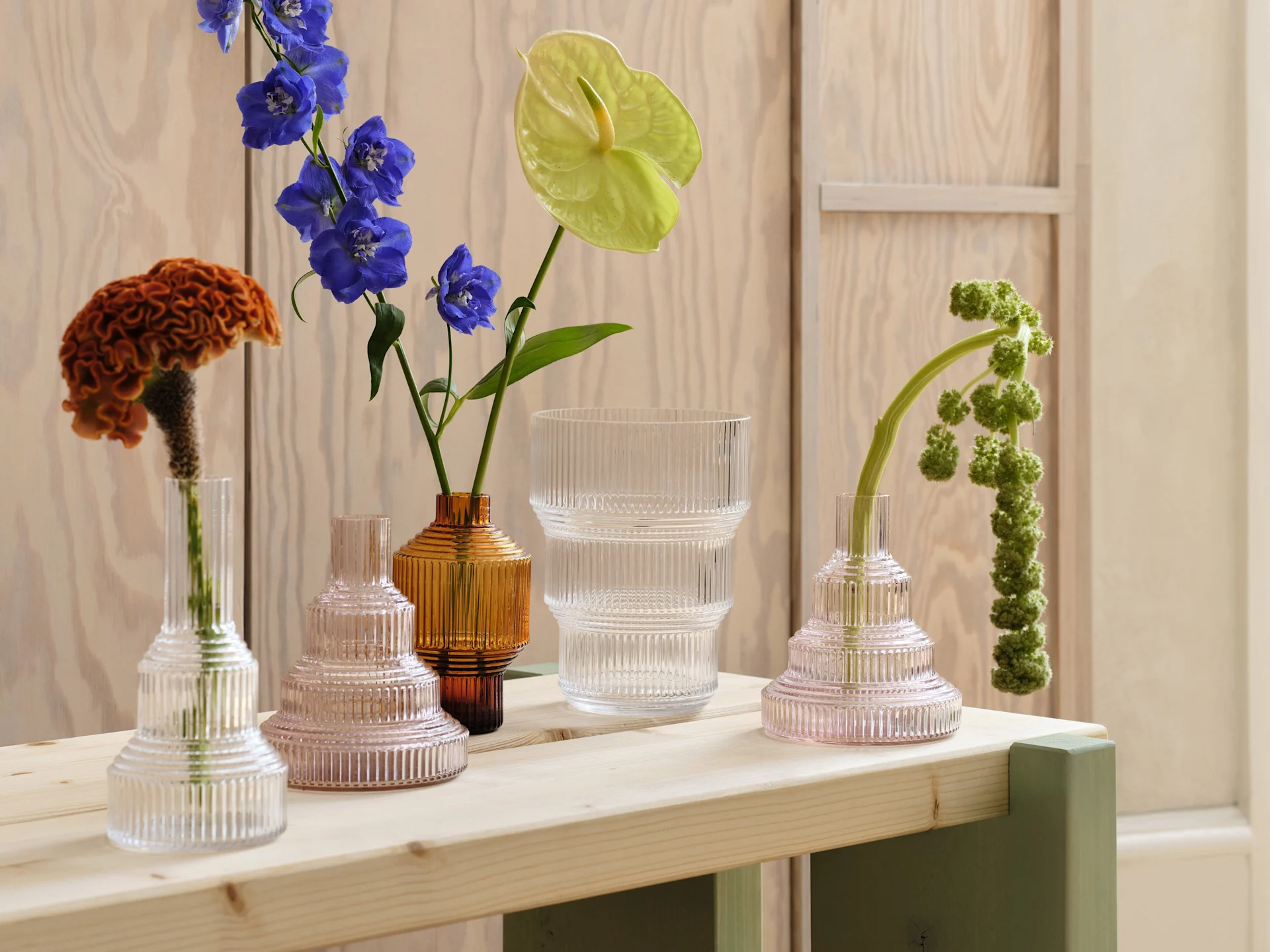 Assortment of ribbed glass vases in clear, pink, and amber with various flowers on a light wood table.
