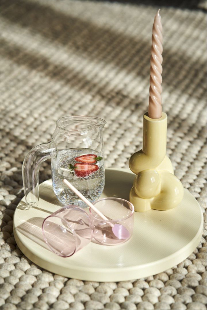 Here you see a collection of HAY products: The Tint drinking glasses in pink, the twist glass straw, the Glass Jug S and the yellow W&S soft candle holder all gathered on a yellow tray. 