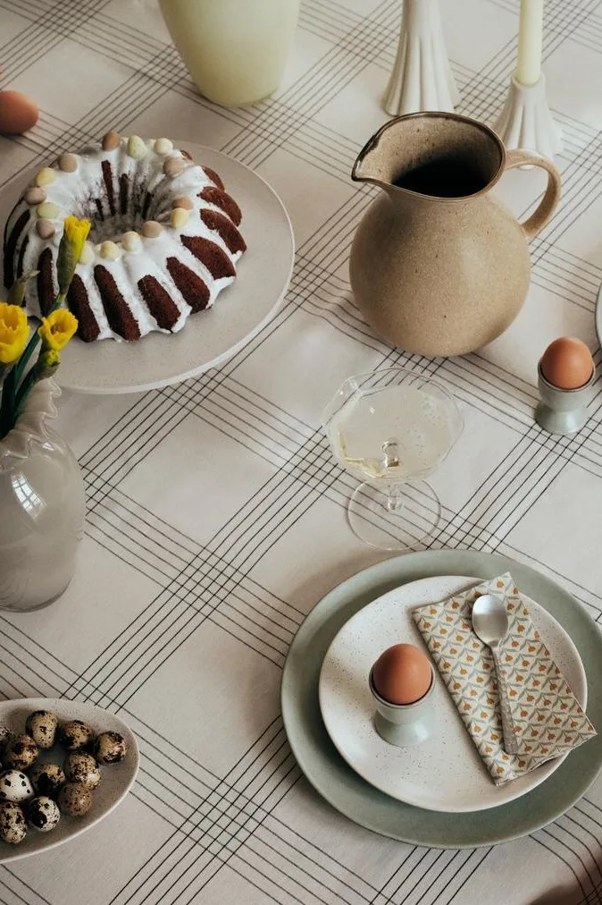 A table set for Easter with a bundt cake, daffodils, eggs, and drinks on a grid tablecloth.
