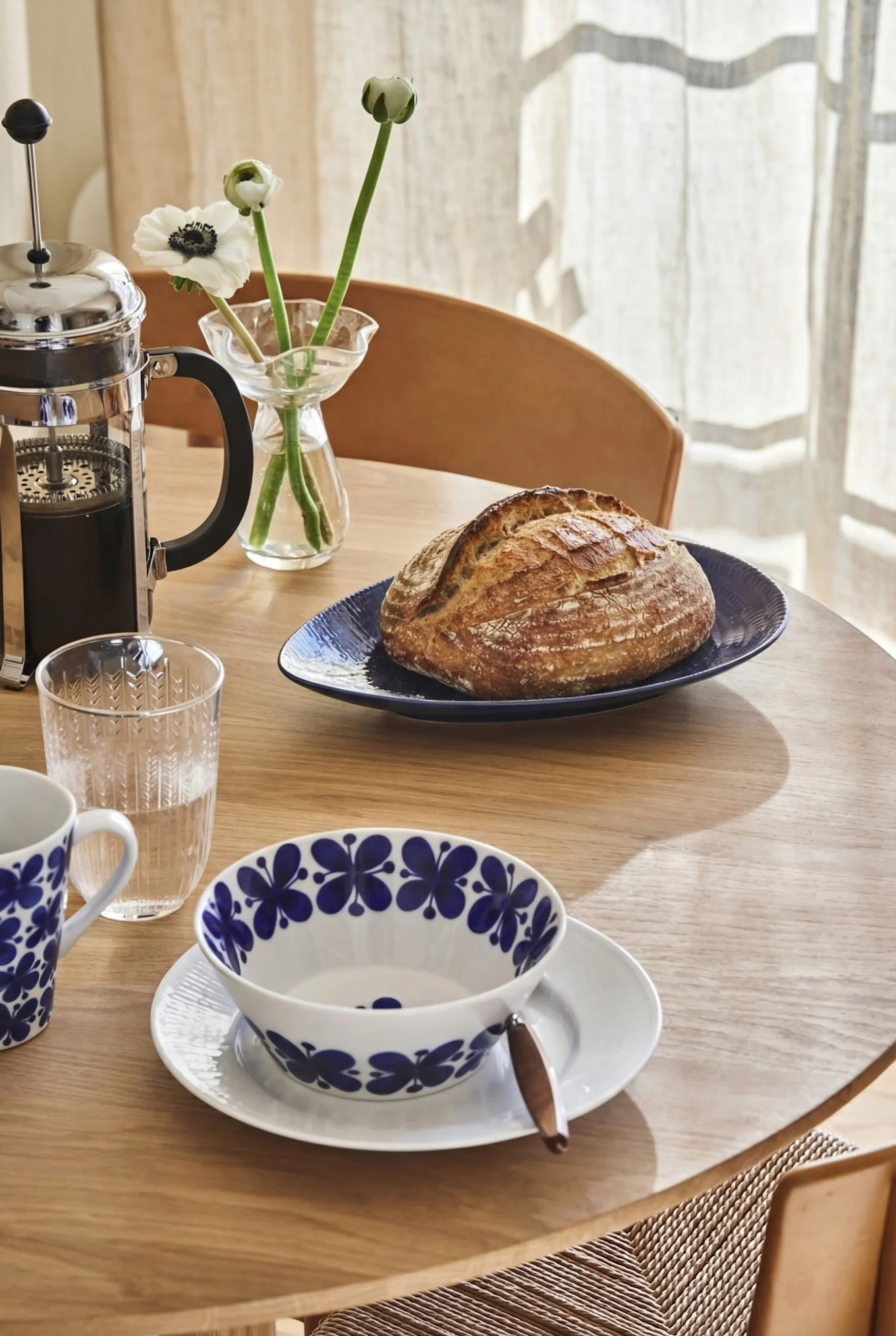 A breakfast table with a French press, rustic bread, water glass, and white and blue patterned ceramic dishes.