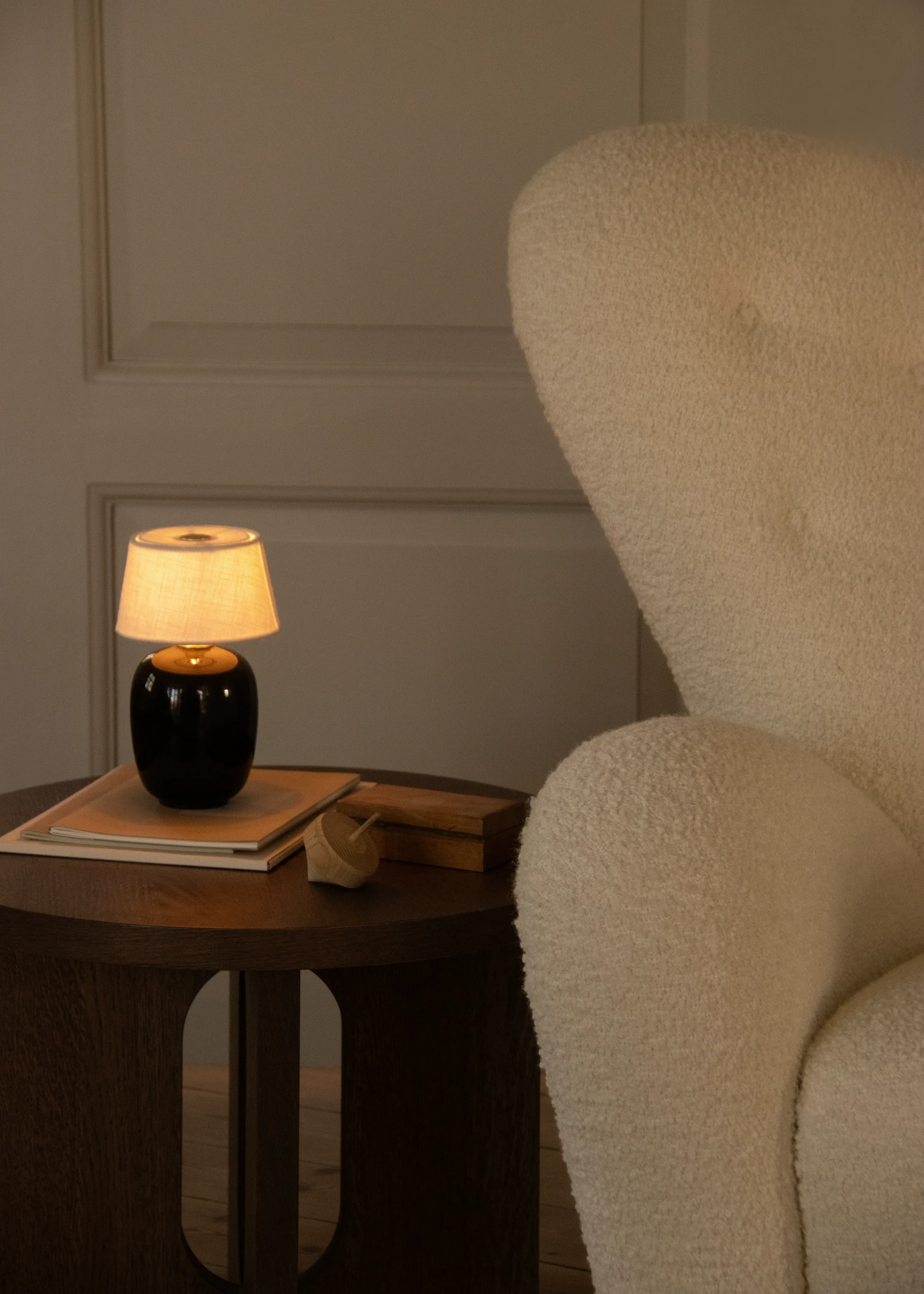 Warmly lit cozy corner with a cream textured armchair and a wooden side table holding a glowing lamp, books, and a wooden toy.