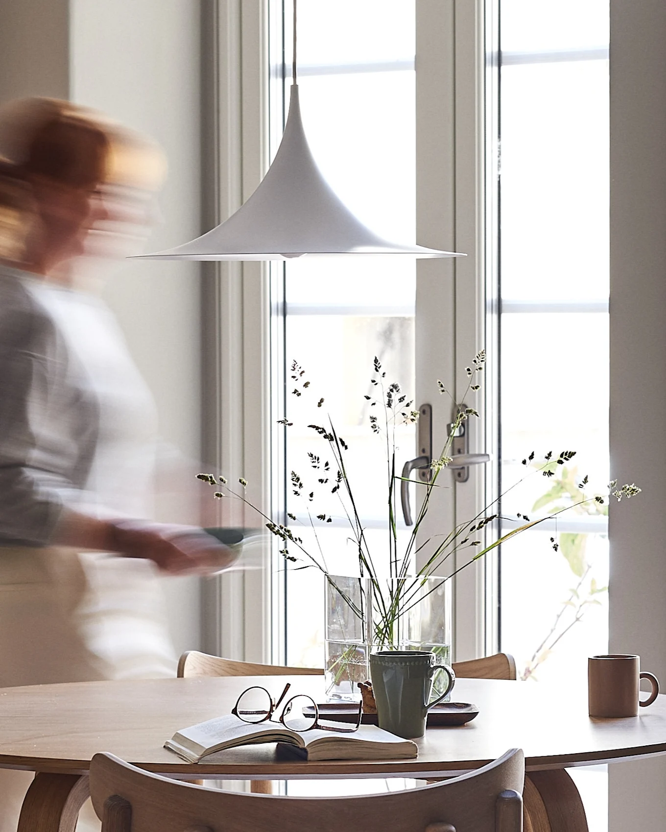 A white pendant lamp hangs over a light wooden table with a book, glasses, and mugs. A blurry person walks by a sunlit window.