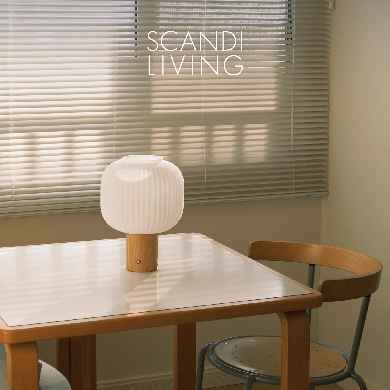 A white ribbed table lamp with a wooden base on a white-topped table and a chair, in front of window blinds. Text: SCANDI LIVING.