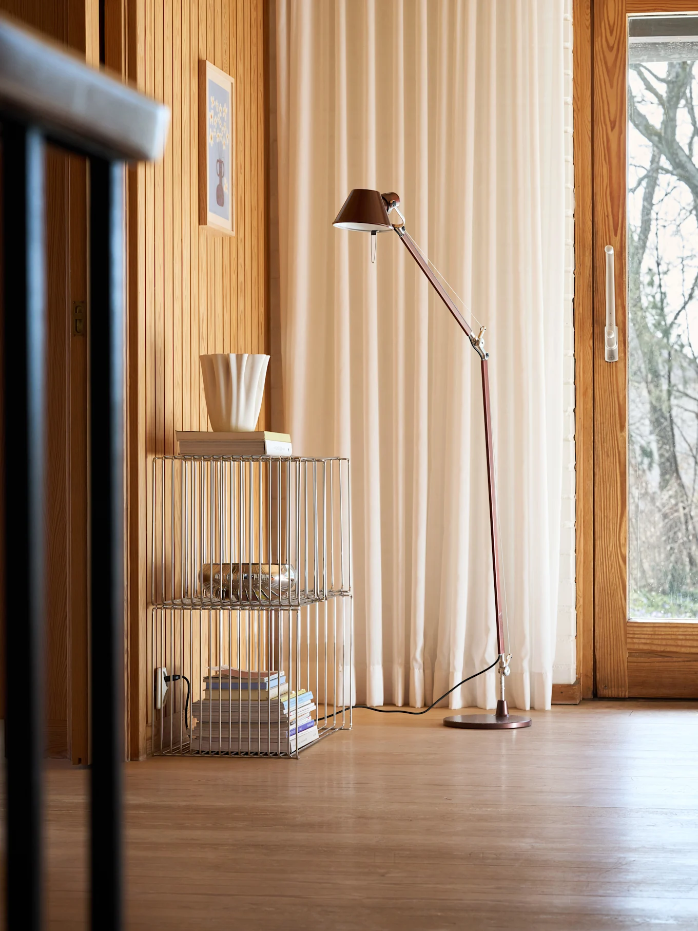 Room with wooden walls and floor, a red floor lamp, a chrome shelf with books and a fluted white vase, and a window with light curtains.