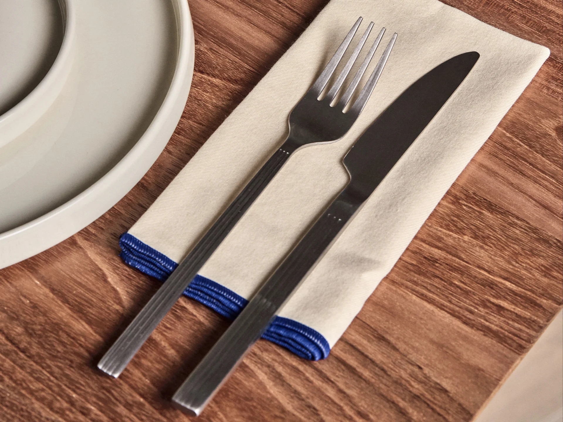 Fork and knife on a cream napkin with a blue border, next to a white plate on a wooden table.