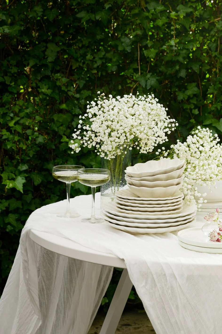 Outdoor table set with white dishes, champagne glasses, and baby's breath flowers against green foliage.