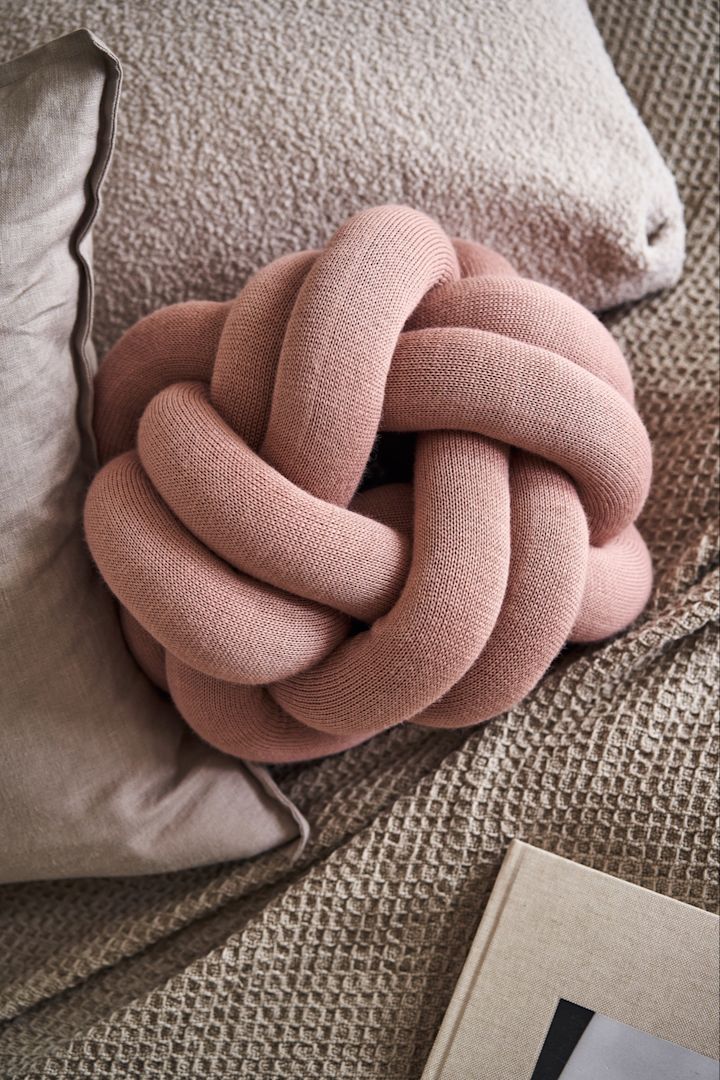 The Knot Cushion small in dusty pink sitting on a bed with beige and grey bed sheets. 