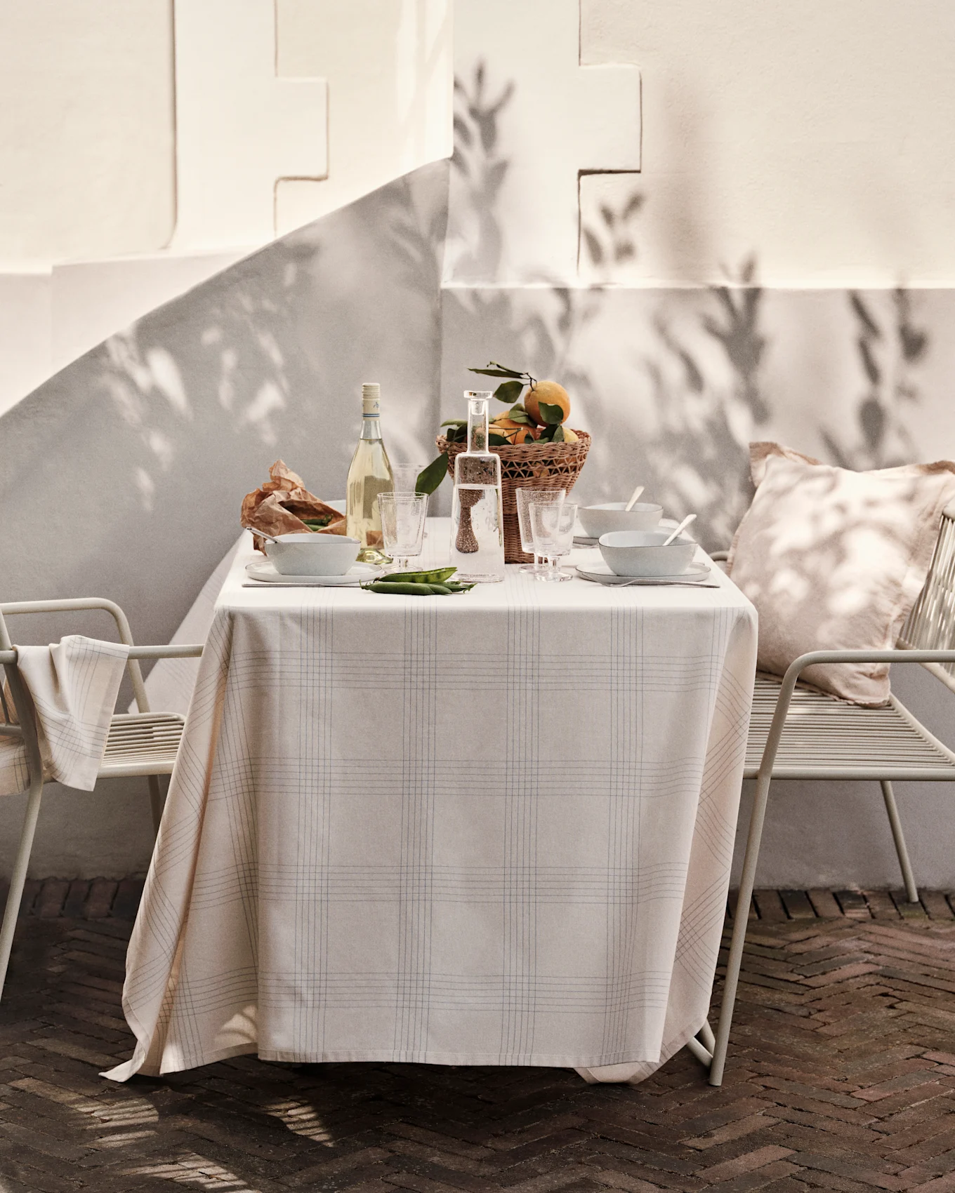 Outdoor table set with plaid tablecloth, wine, water, bowls, basket of oranges, and peas on a sunny patio.