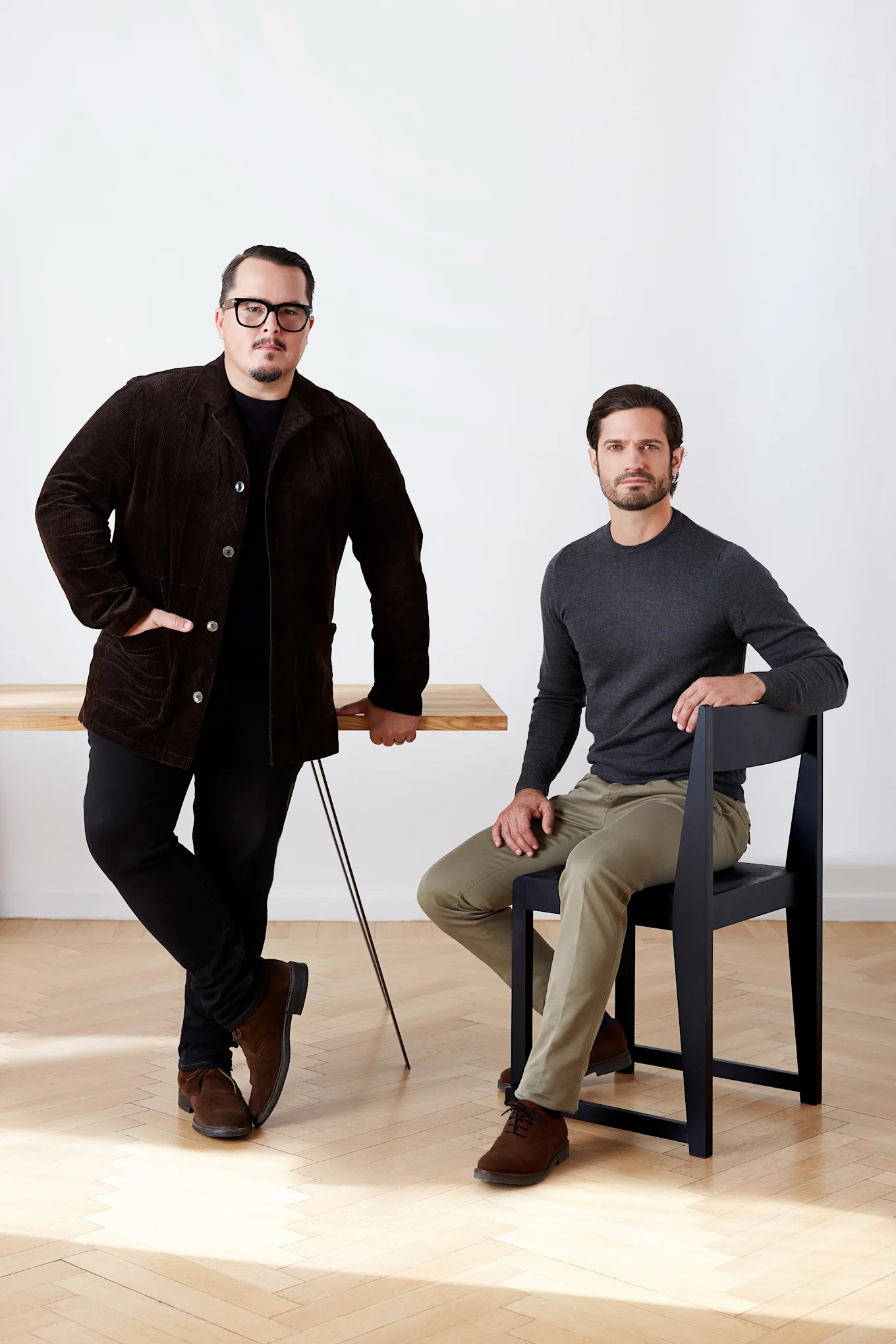 Two men, one standing by a wooden table in a brown corduroy jacket, the other sitting on a black chair in a gray shirt.