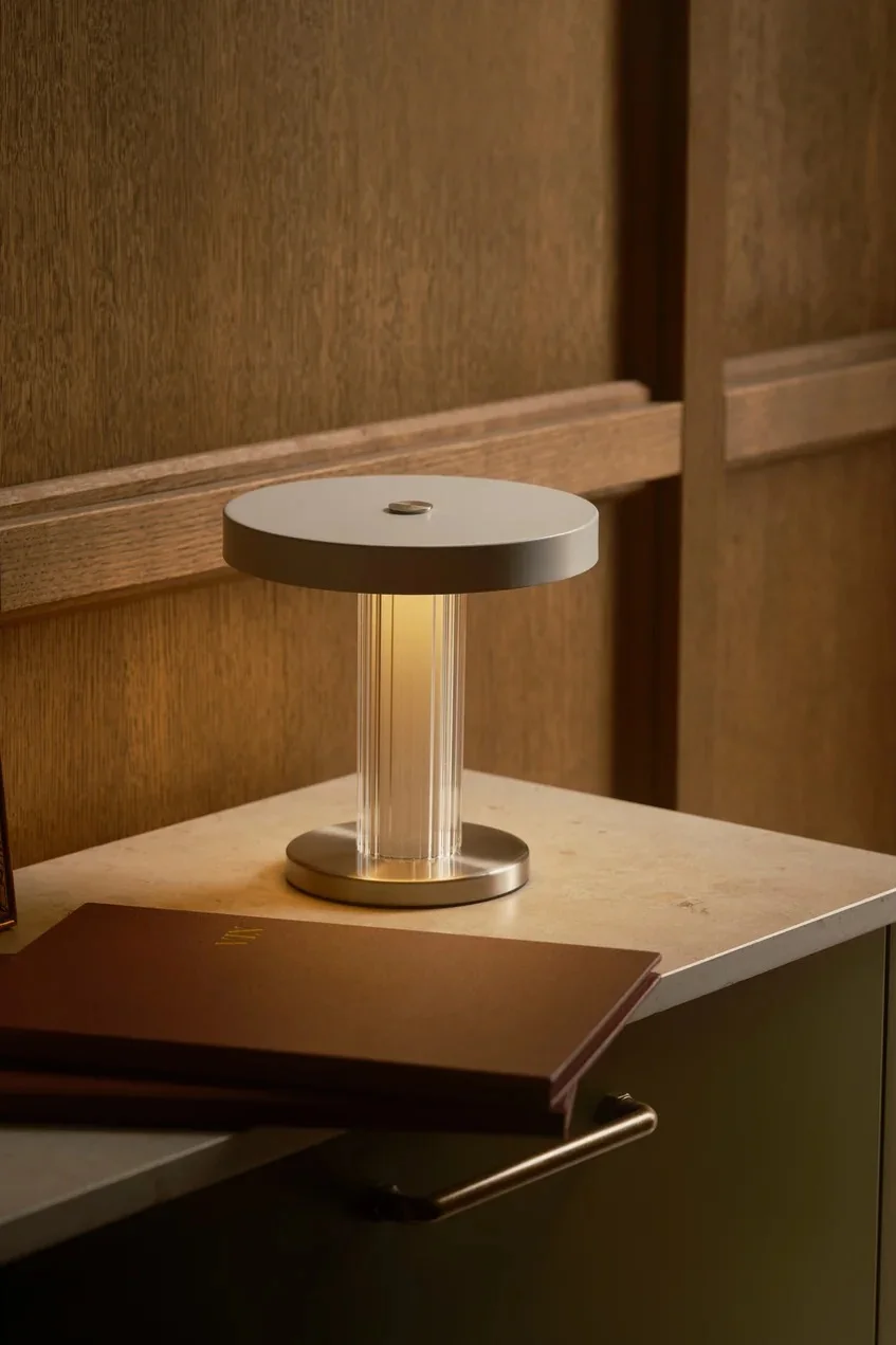A modern table lamp with a ribbed, transparent column and circular base and top, emitting a warm glow on a countertop next to a brown booklet.