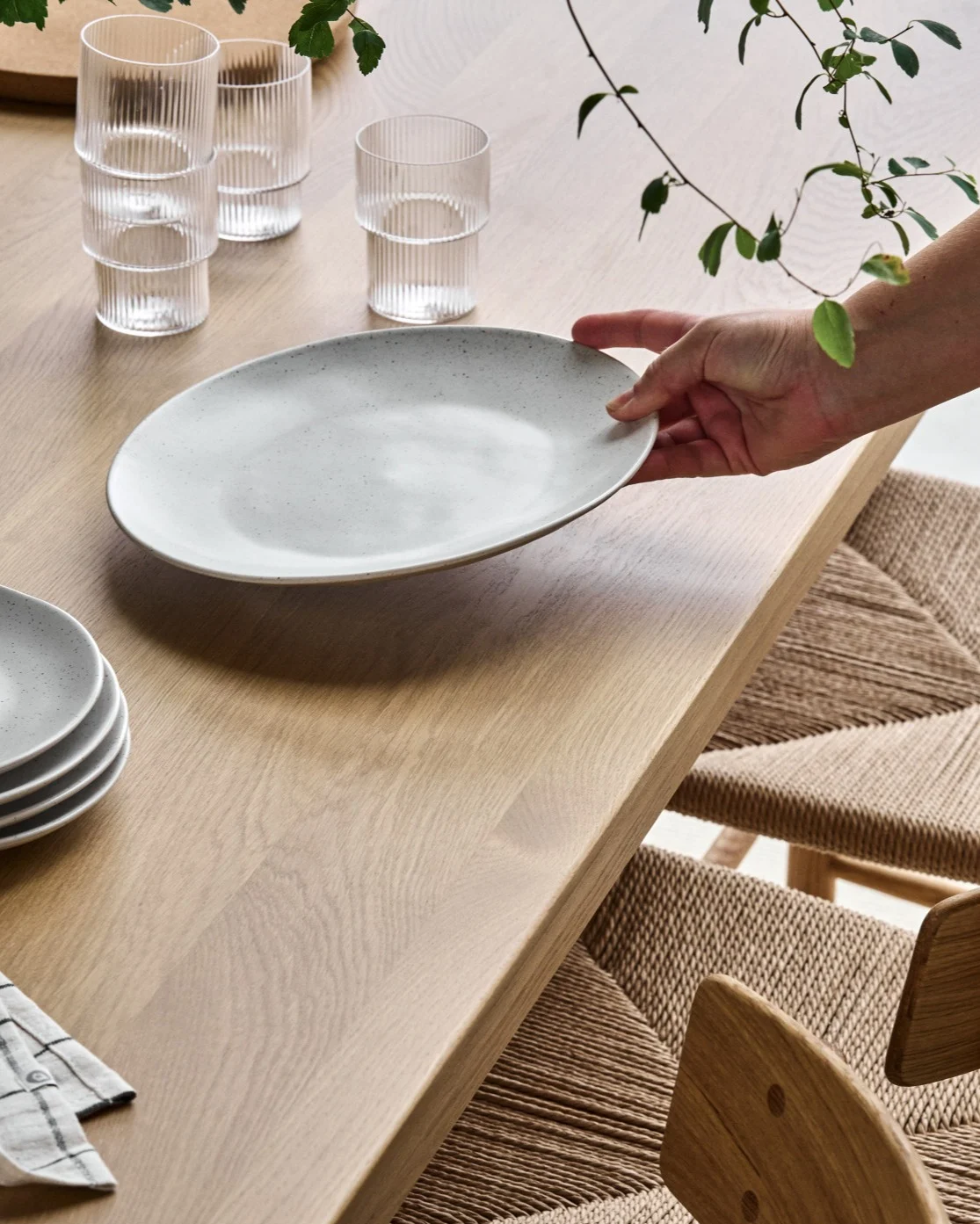 A person setting a light grey speckled oval plate on a wooden dining table, with matching chairs and ribbed glasses.