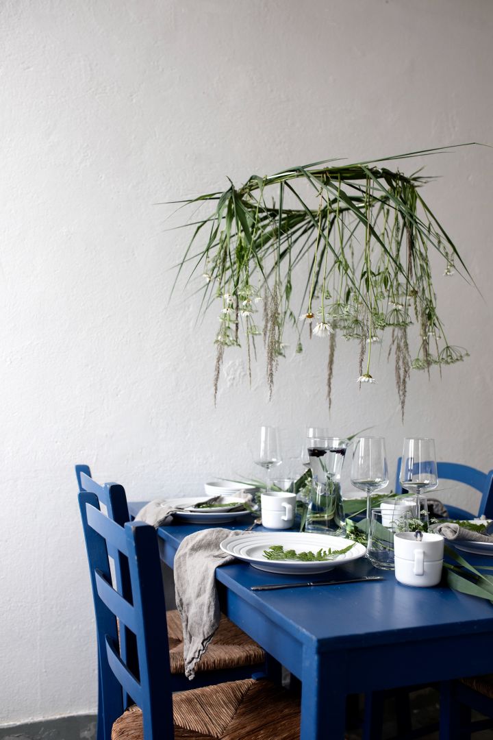 A blue dining table  set with the NJRD Lines porcelain set, with white walls with greenery hanging over the table make the perfect Mediterranean themed decor.