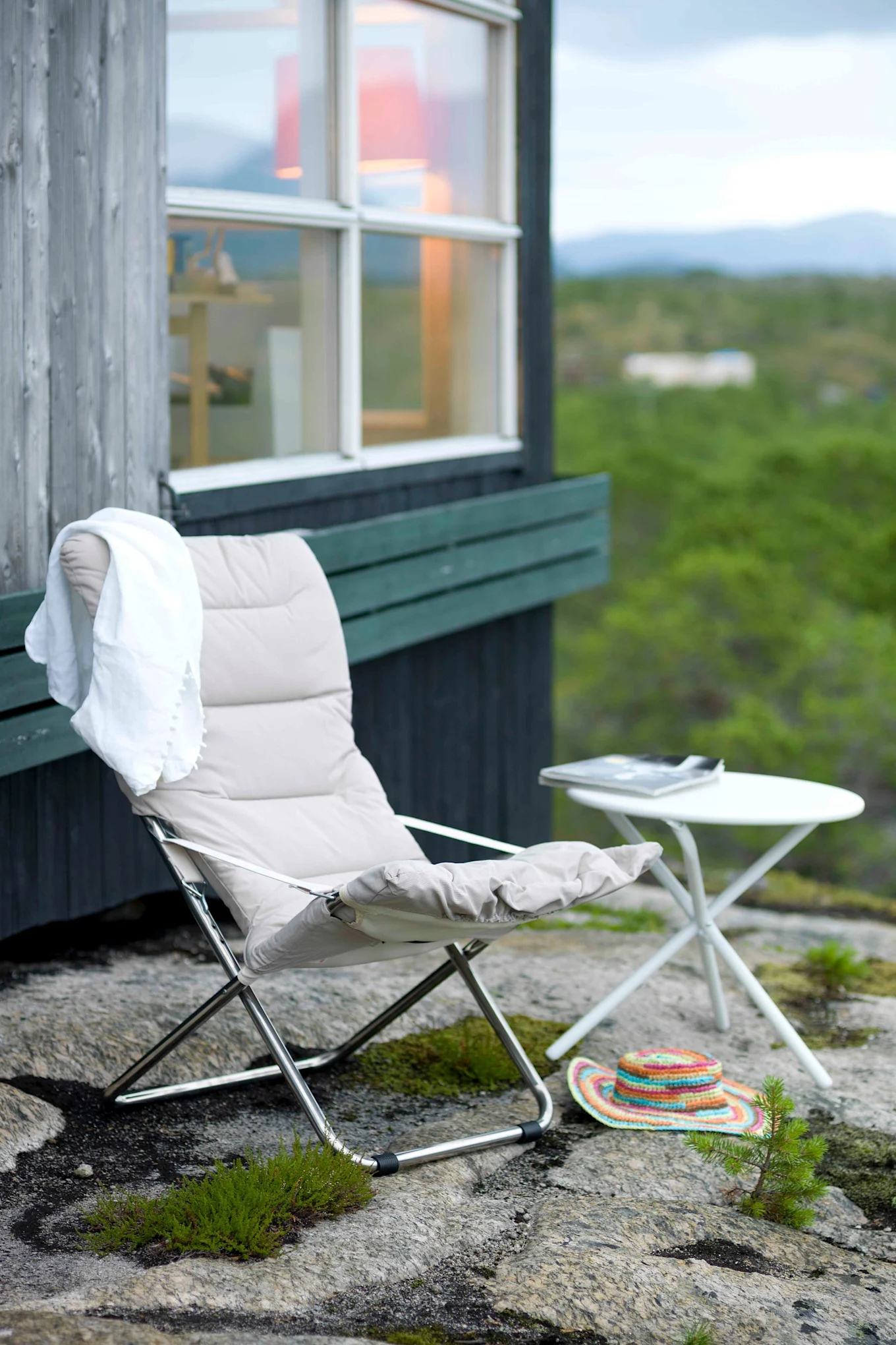 A padded folding chair with a white towel, a small white table, and a colorful hat on rocky ground outside a cabin.