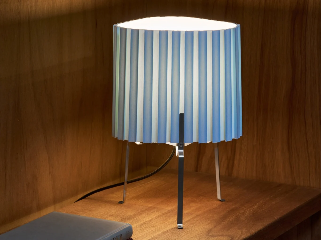 Table lamp with pleated blue and white striped shade on a wooden shelf, illuminating the area.