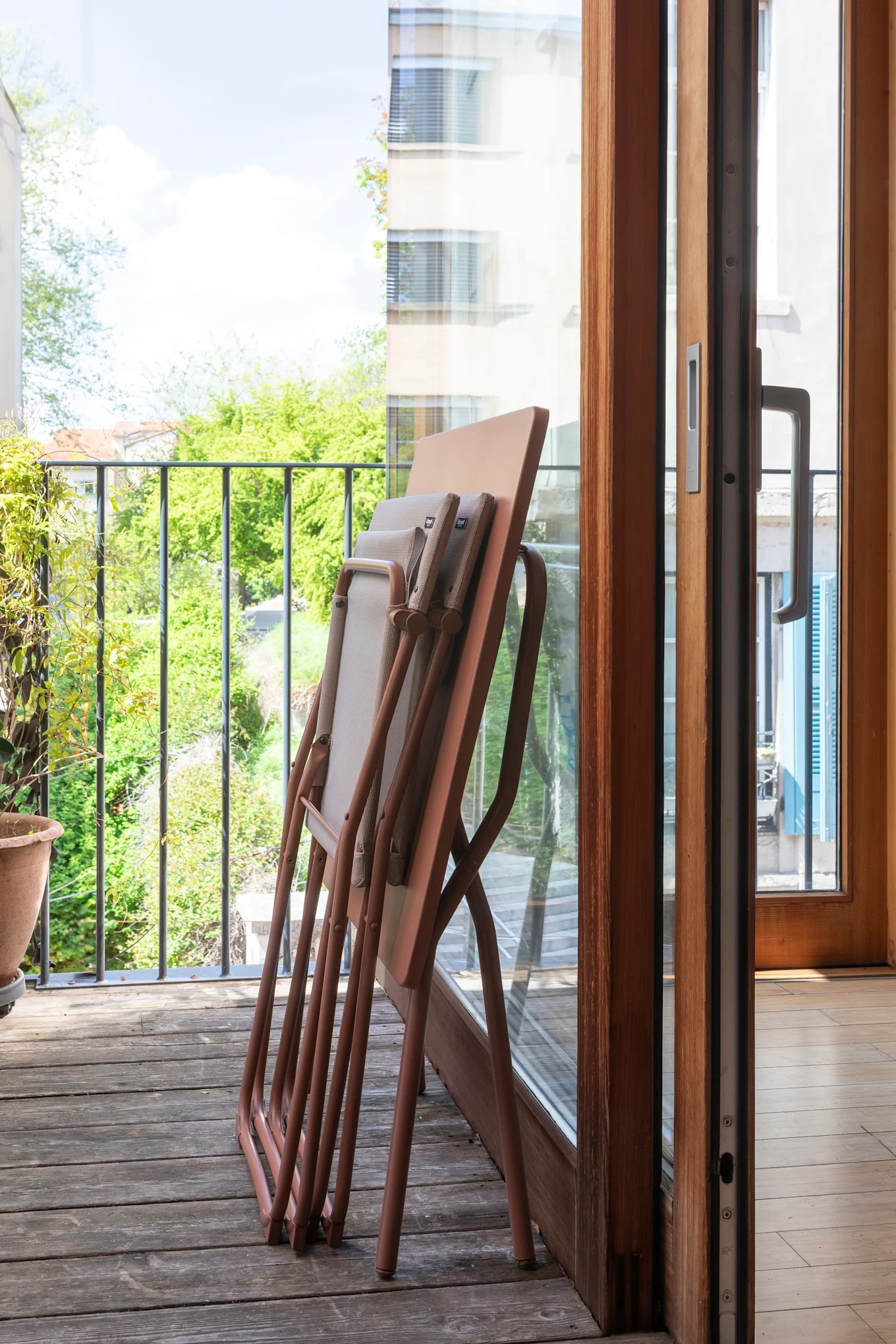 Folded chairs stacked on a wooden balcony floor next to a glass door, overlooking green trees.