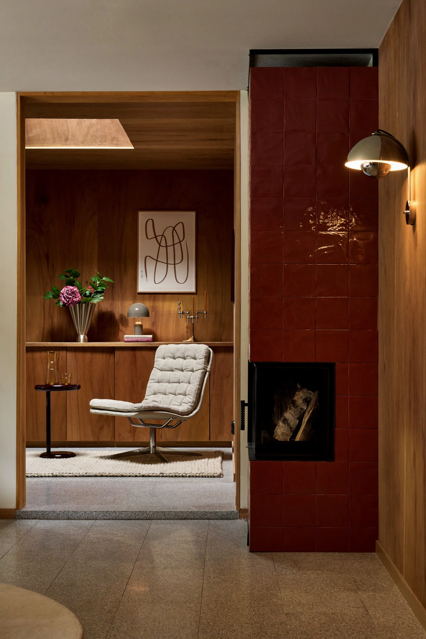 A cozy interior featuring a textured lounge chair, wood-paneled walls, a red-tiled fireplace, and a gold wall sconce.