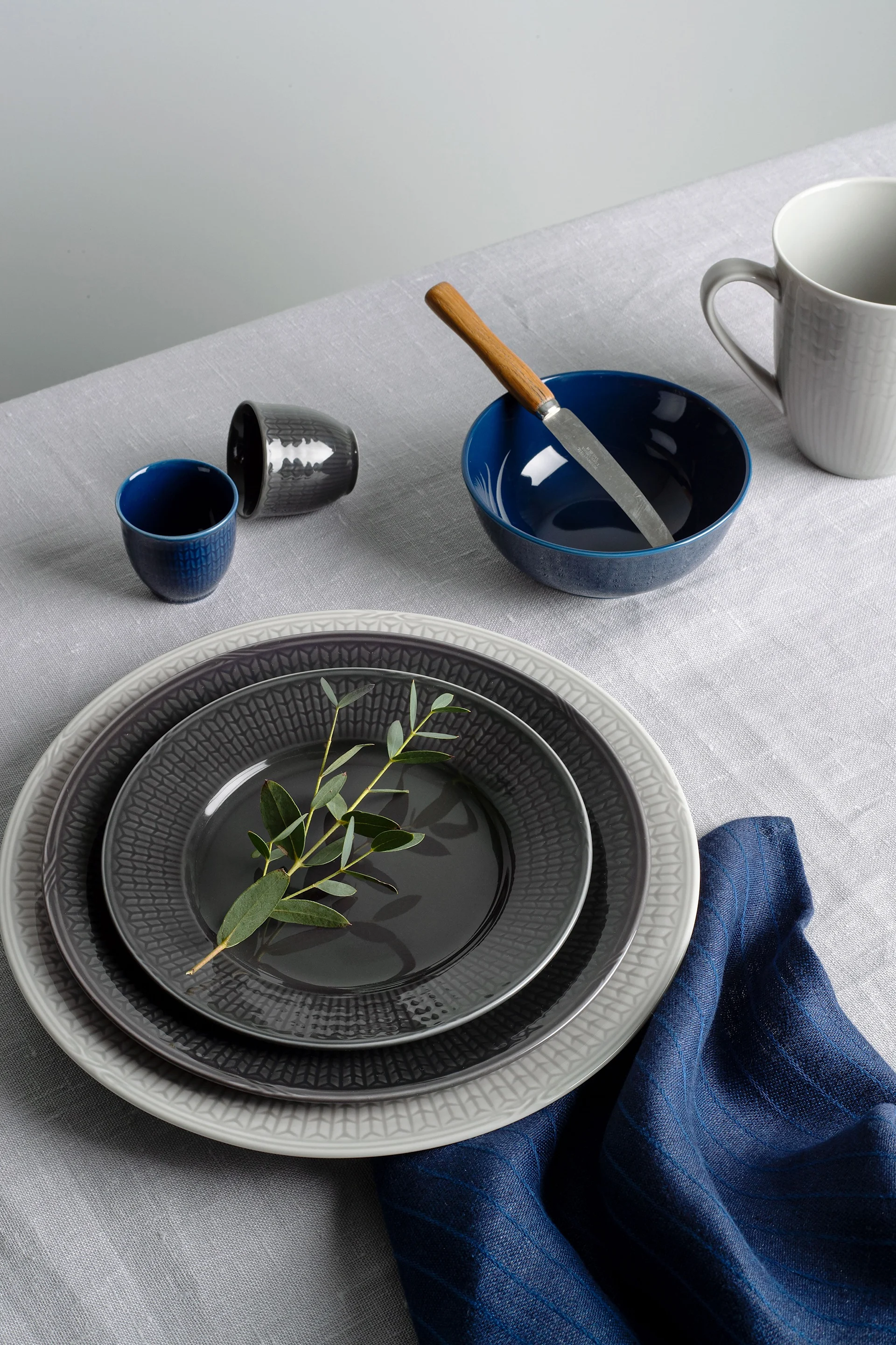 A light grey table setting with stacked dark grey and light grey ceramic plates, blue and grey bowls, cups, a knife, and a blue napkin.