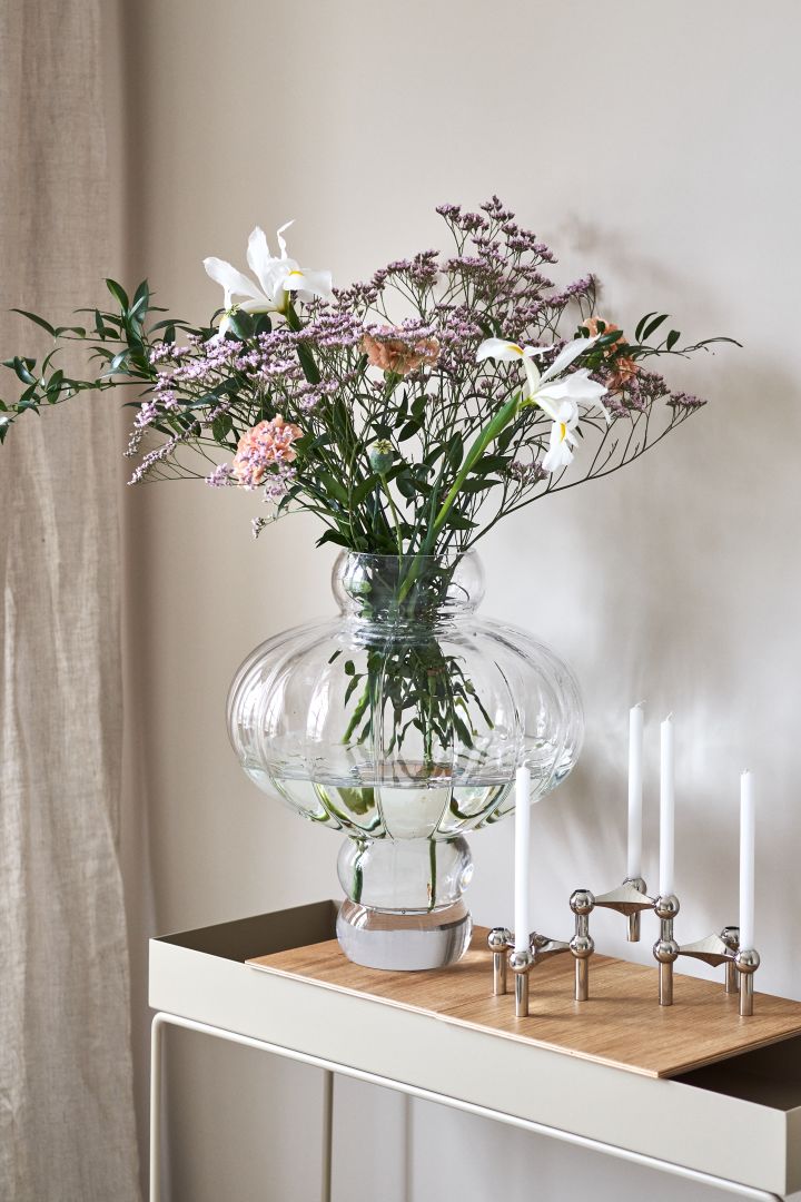 The round and elegant Balloon vase in clear glass from Louise Roe Copenhagen is a unique large vase next to the Stoff candle holder on the Plant Box from Ferm Living. 