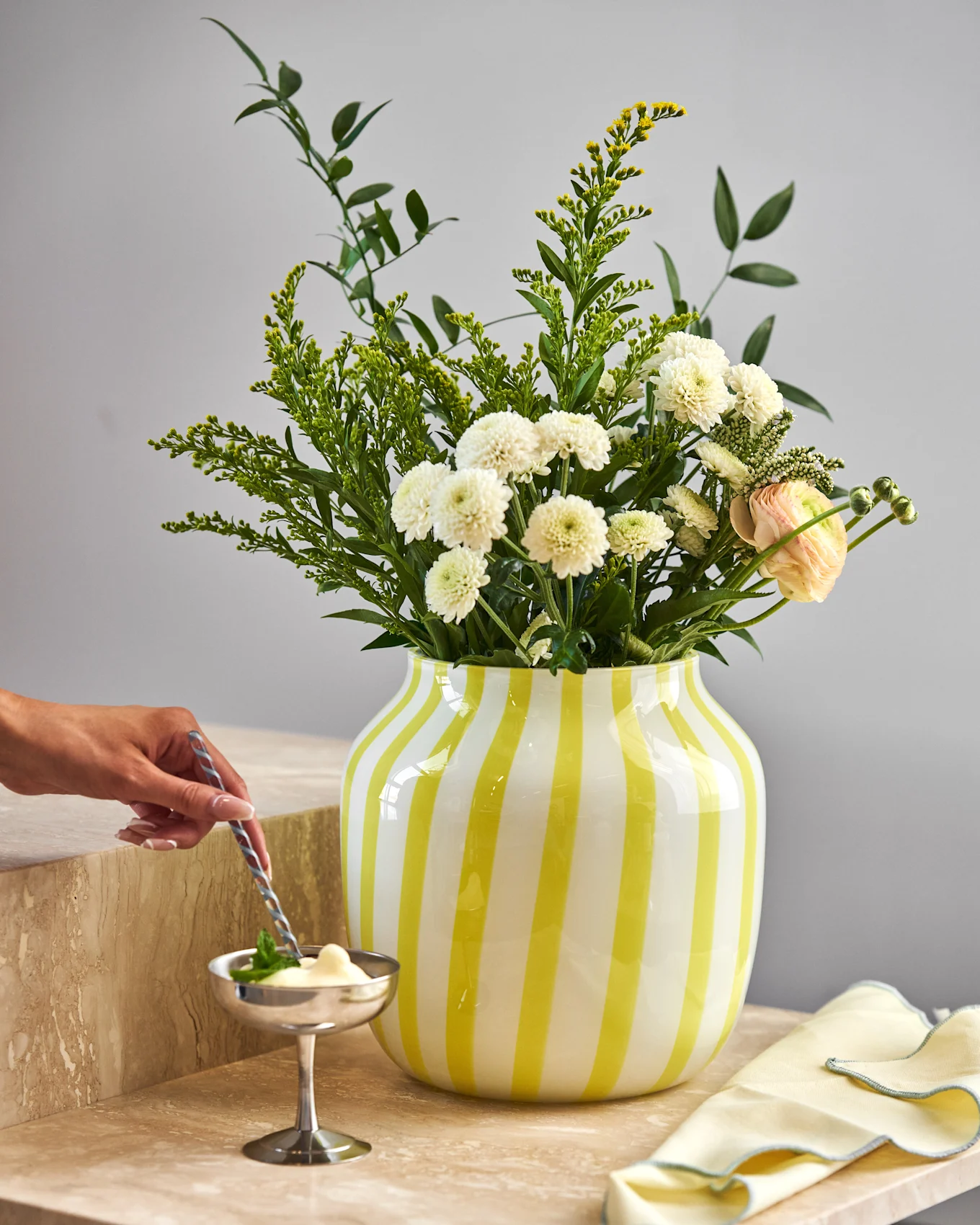Hand stirs dessert in silver bowl on marble. Yellow striped vase holds white chrysanthemums, yellow flowers, and pink ranunculus.