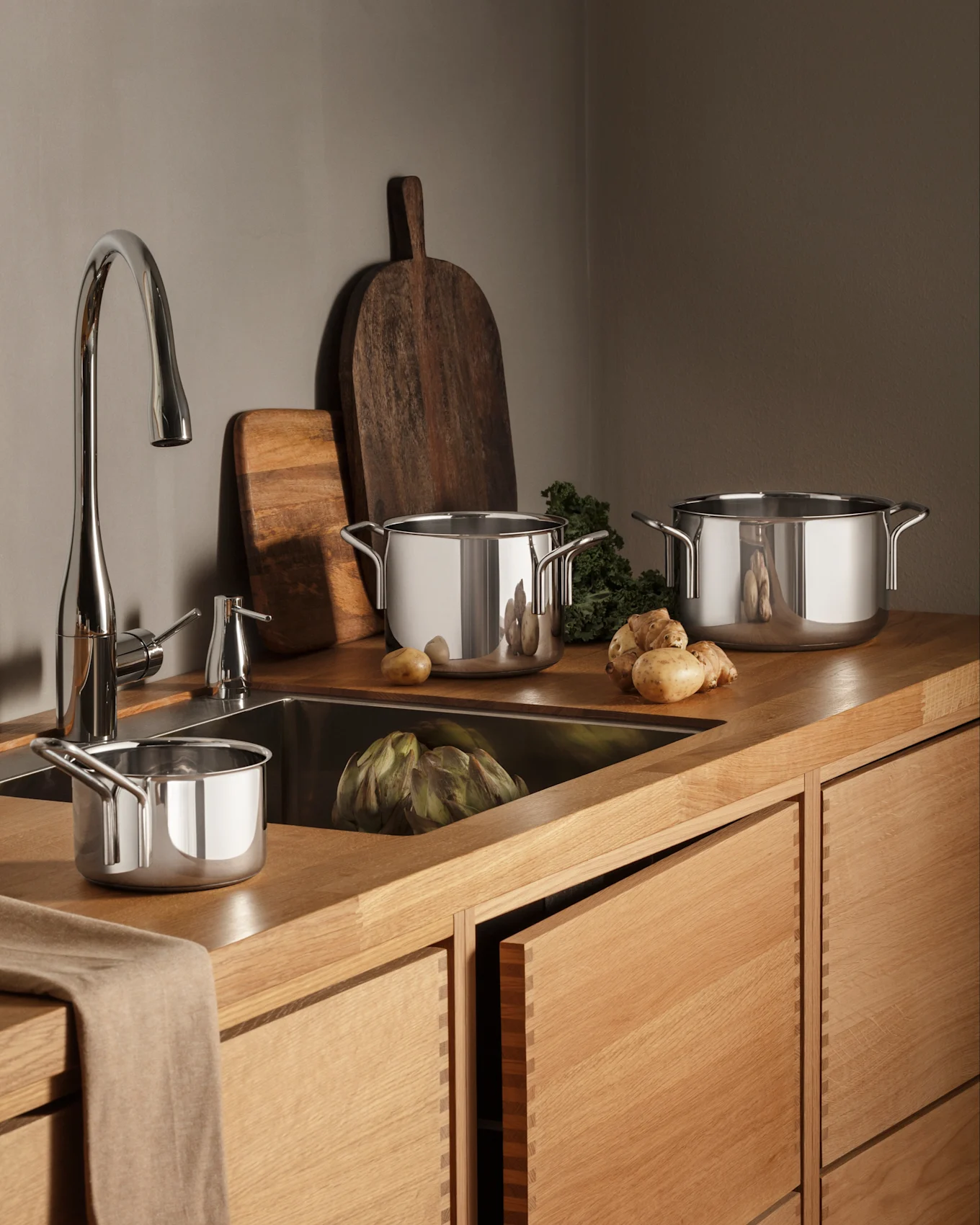 A modern kitchen with wooden counters and cabinets, a chrome faucet, and stainless steel pots, fresh produce including artichoke and potatoes.