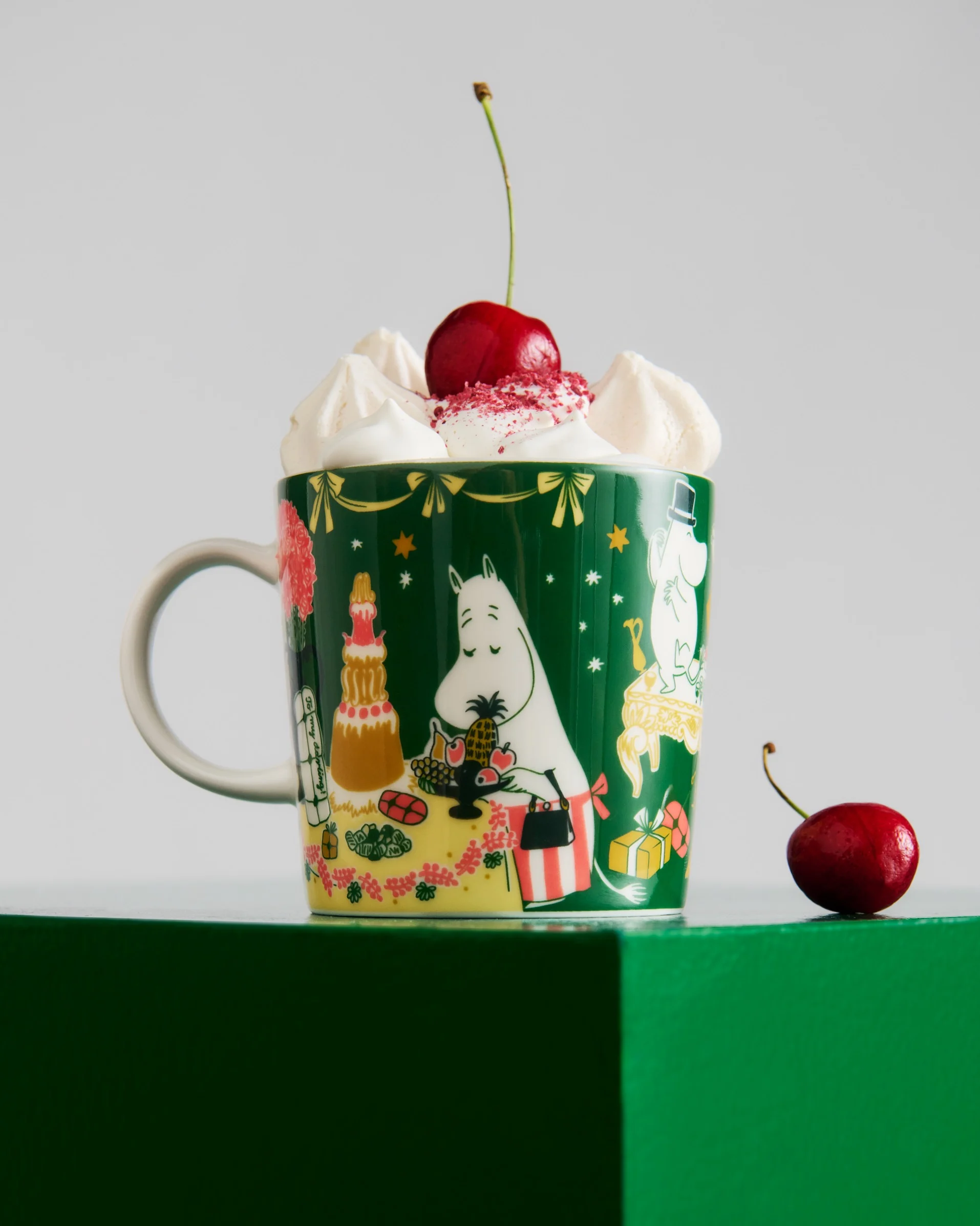 Dark green mug with Moomin characters, topped with whipped cream, red sprinkles, and a cherry. Another cherry sits on the green surface.