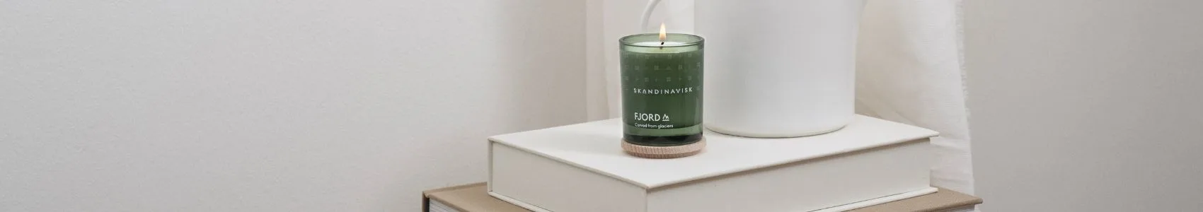 A book titled 'NOTES FROM THE FELLS' on a grey blanket next to two Scandinavian-style candles, one black and burning, one brown with a wooden lid.