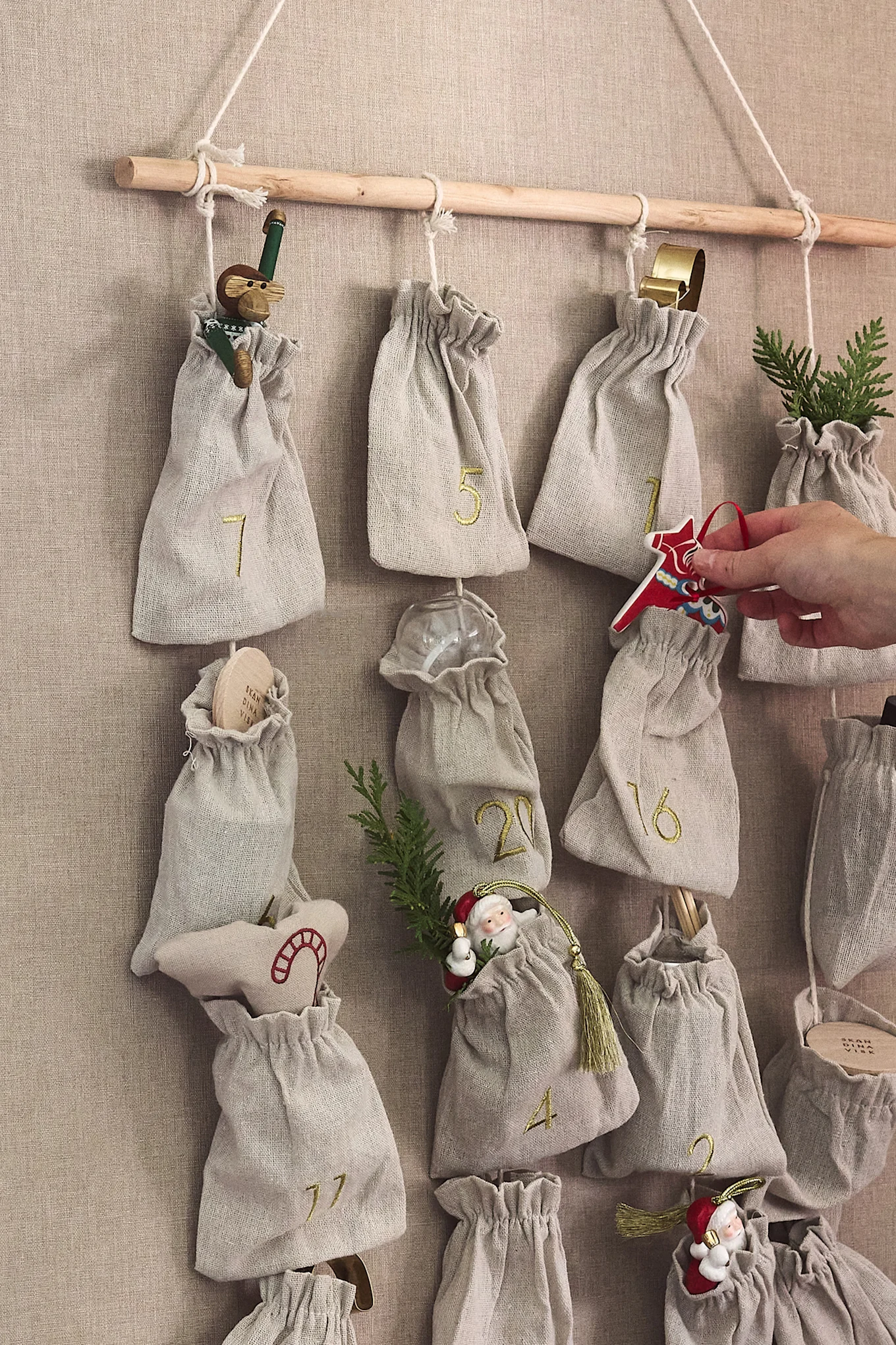 Fabric advent calendar with numbered bags hanging from a wooden rod, containing various Christmas ornaments; a hand places a red Dala horse.