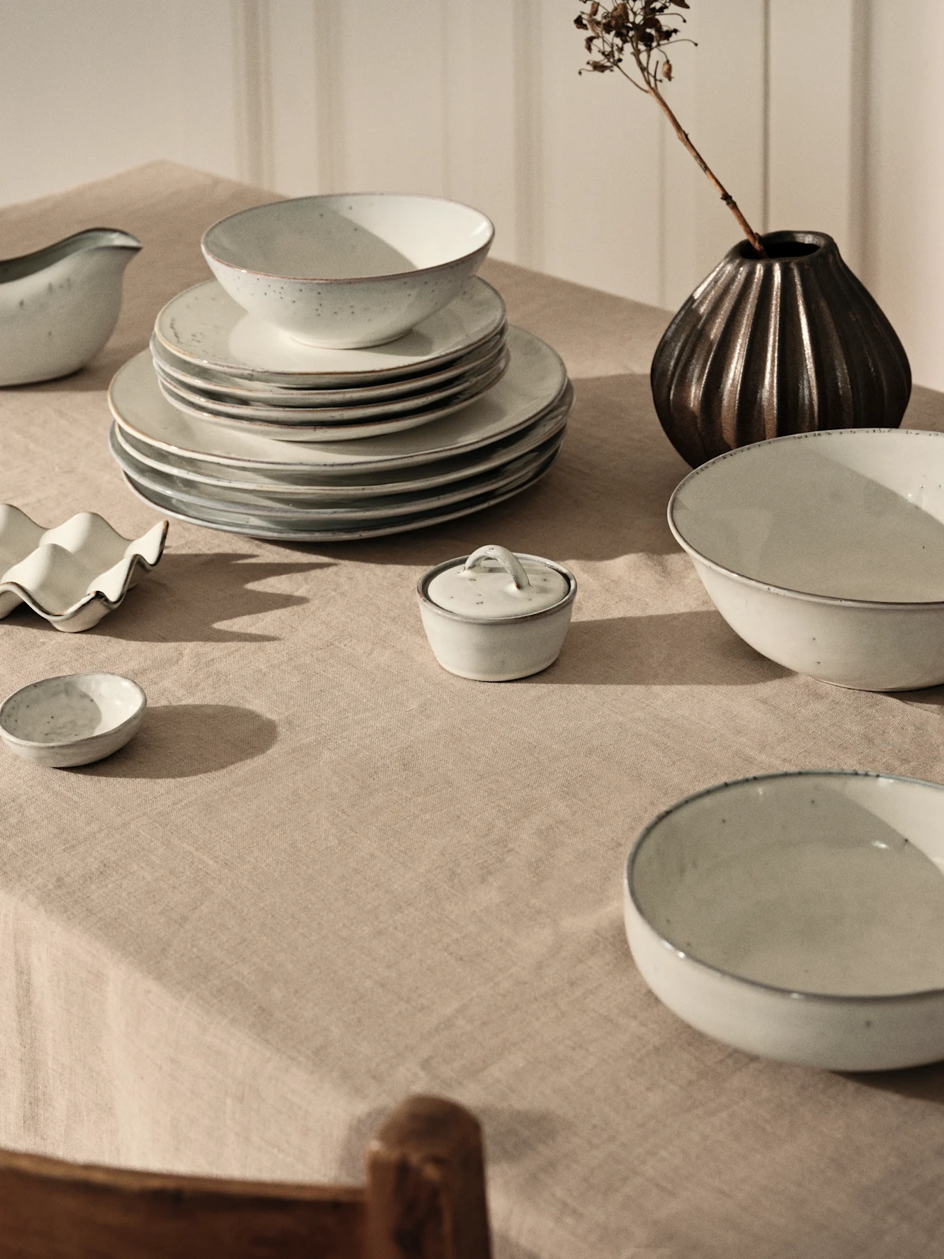 Light grey ceramic dinnerware set with stacked plates and bowls on a beige linen tablecloth, alongside a dark vase with a dried branch.