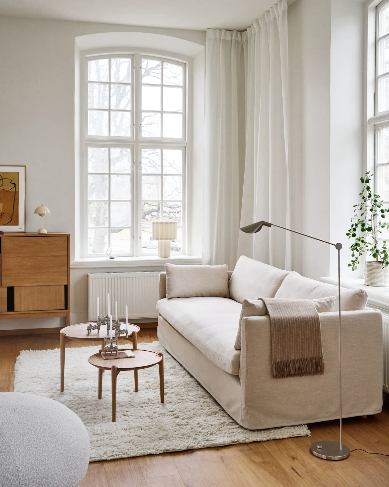 A bright, minimalist living room with a light beige sofa, two round wooden coffee tables with candles, a shaggy white rug, and a large window.