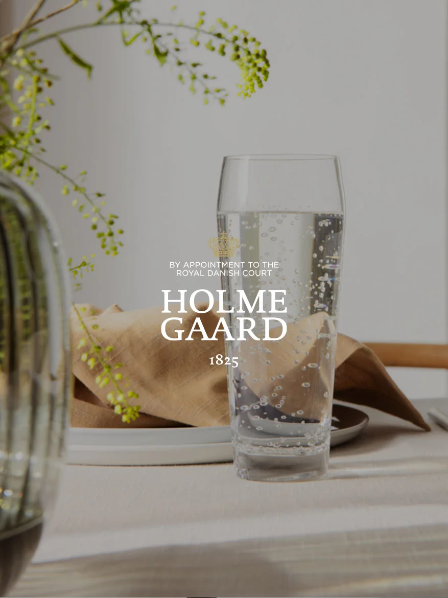 A tall glass of sparkling water next to a brown napkin and a plant, with the Holmegaard logo overlaid.