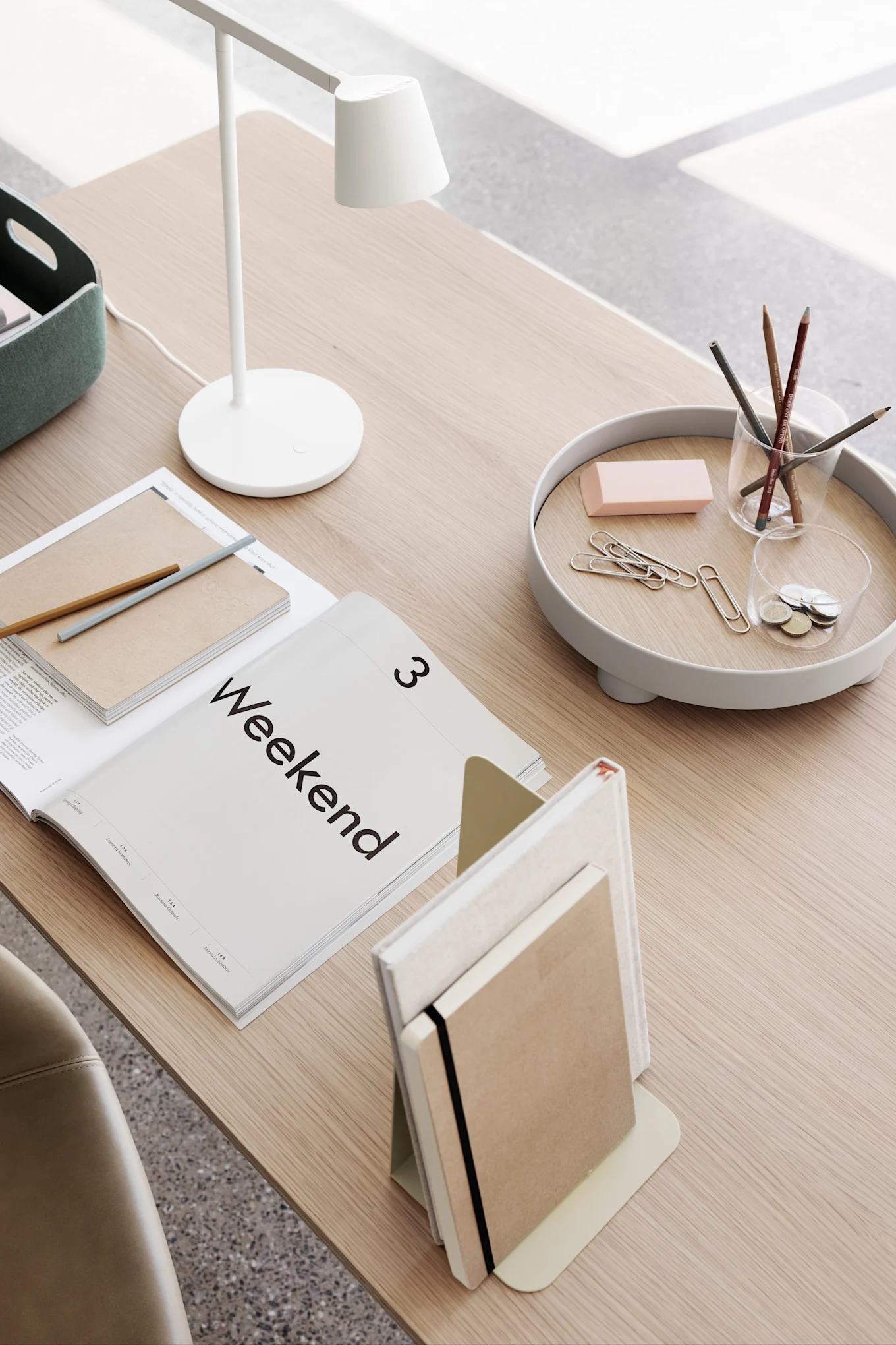 Minimalist desk setup with a white lamp, an open 'Weekend' magazine, notebooks, and desk accessories on a tray.
