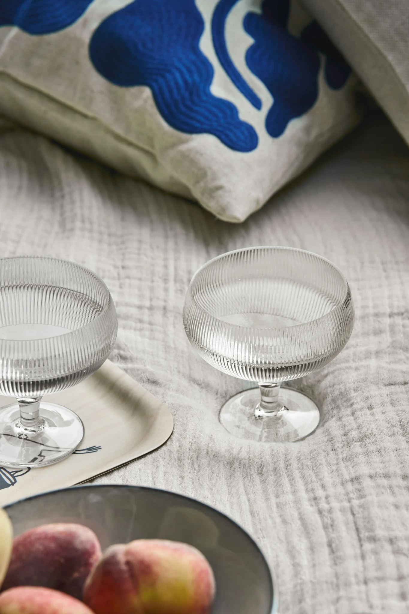 Two ribbed glass coupe glasses on a light grey fabric, with peaches in a bowl and a blue patterned cushion.