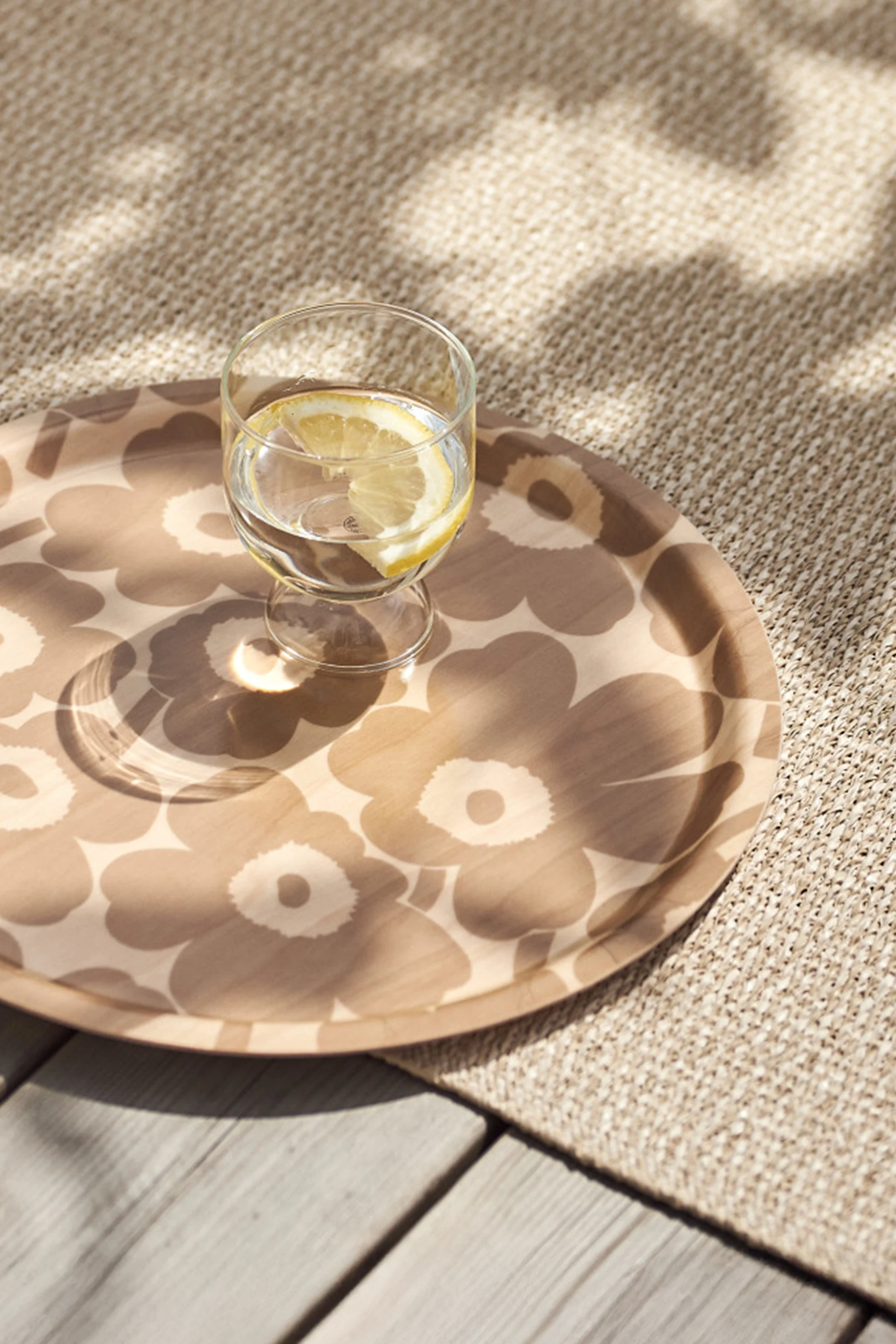 Glass of water with lemon slice on a patterned round tray, placed on a beige woven rug and wooden deck with dappled sunlight.