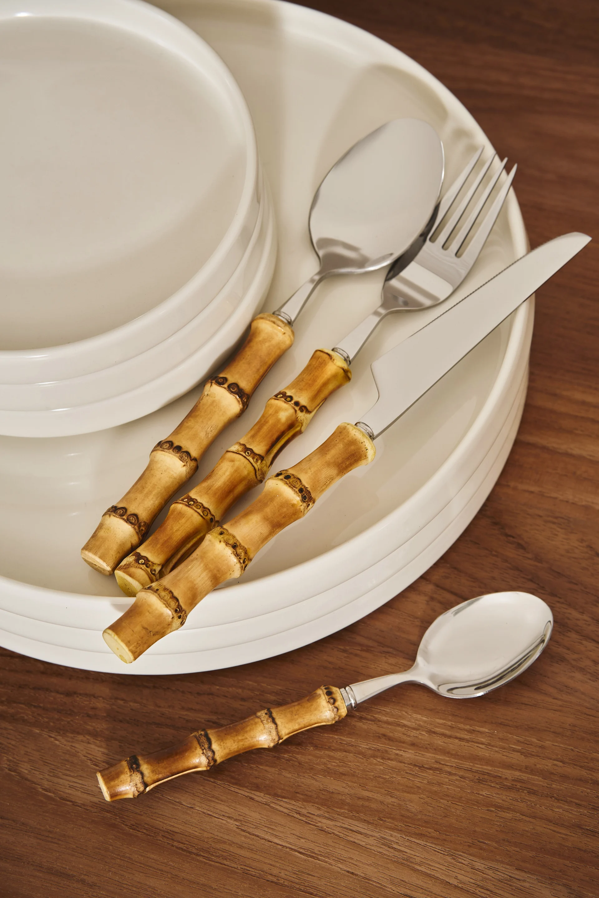 Silver cutlery with bamboo handles on a stack of white plates on a wooden table.